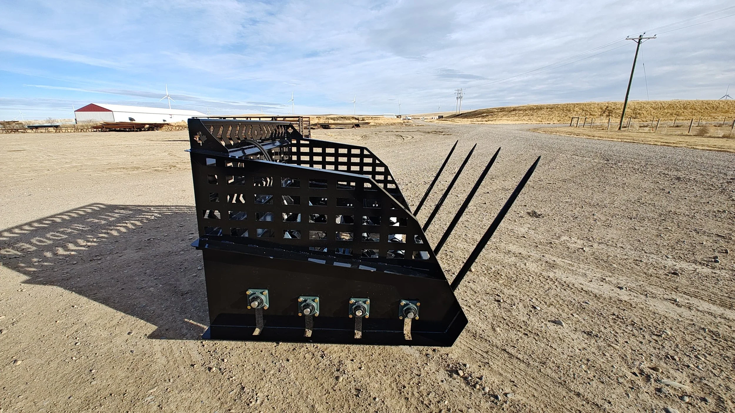 Black agricultural implement with metal spikes in a wide open sandy area with wind turbines and power lines in the background under a cloudy sky.