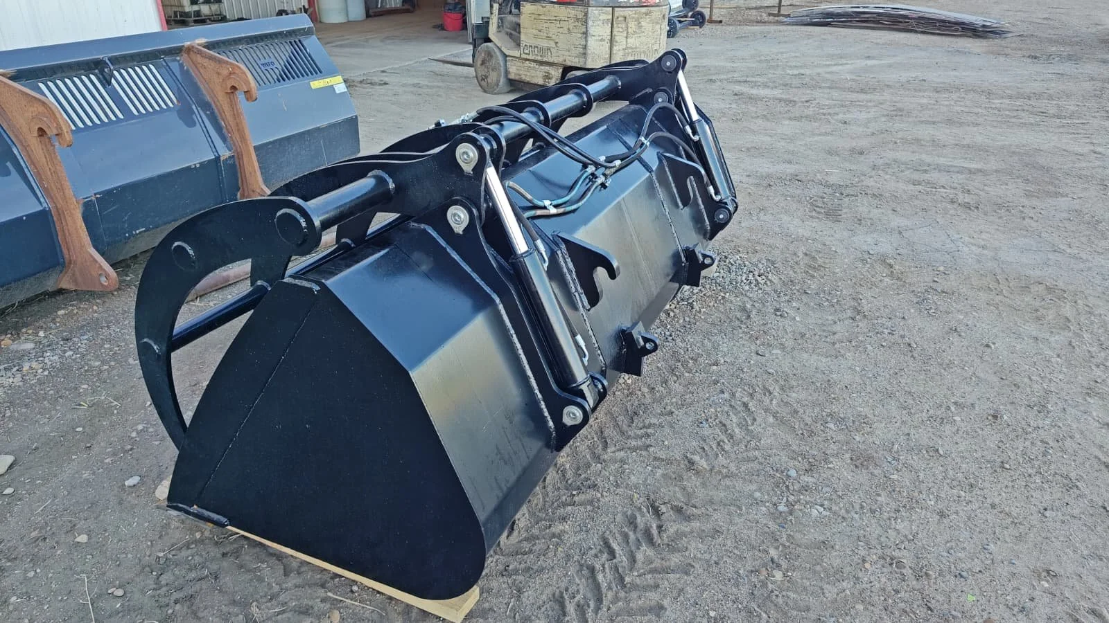 A large black industrial bucket attachment for construction equipment, resting on a dirt ground outdoors.
