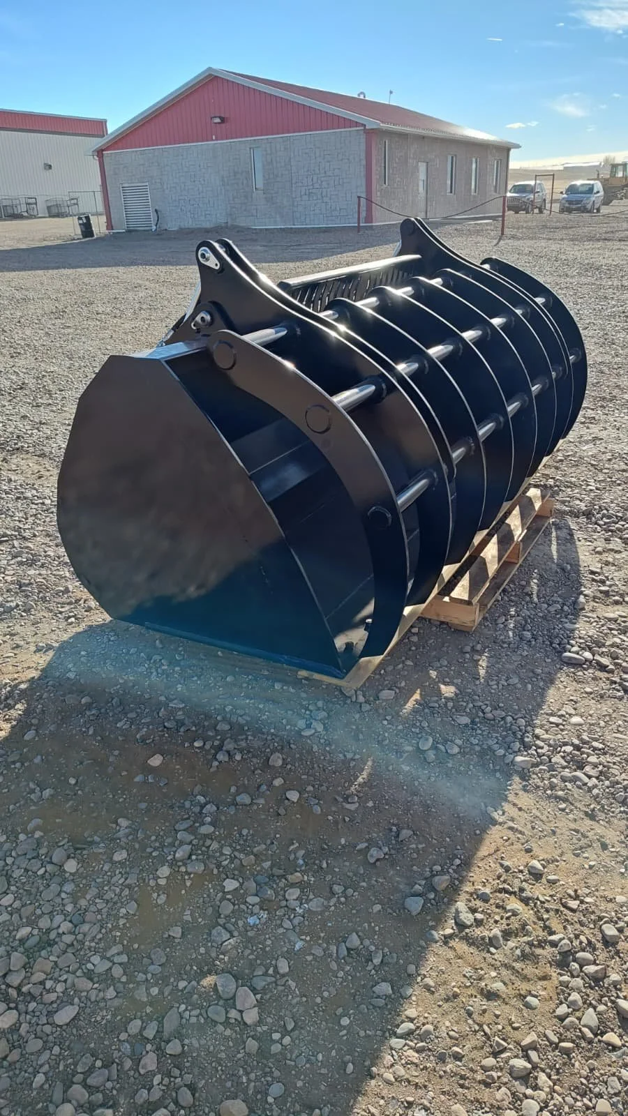 A large black metal scoop attachment for heavy equipment, resting on a wooden pallet outdoors with buildings and vehicles in the background.