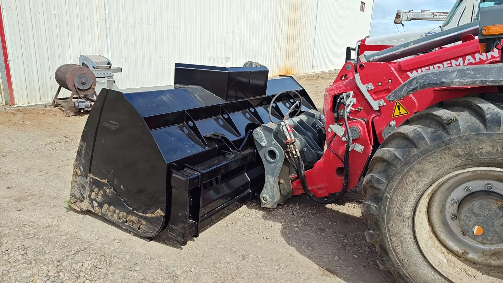 Close-up of a red tractor with a black attachment for clearing land or snow, parked on a dirt surface outside a building.