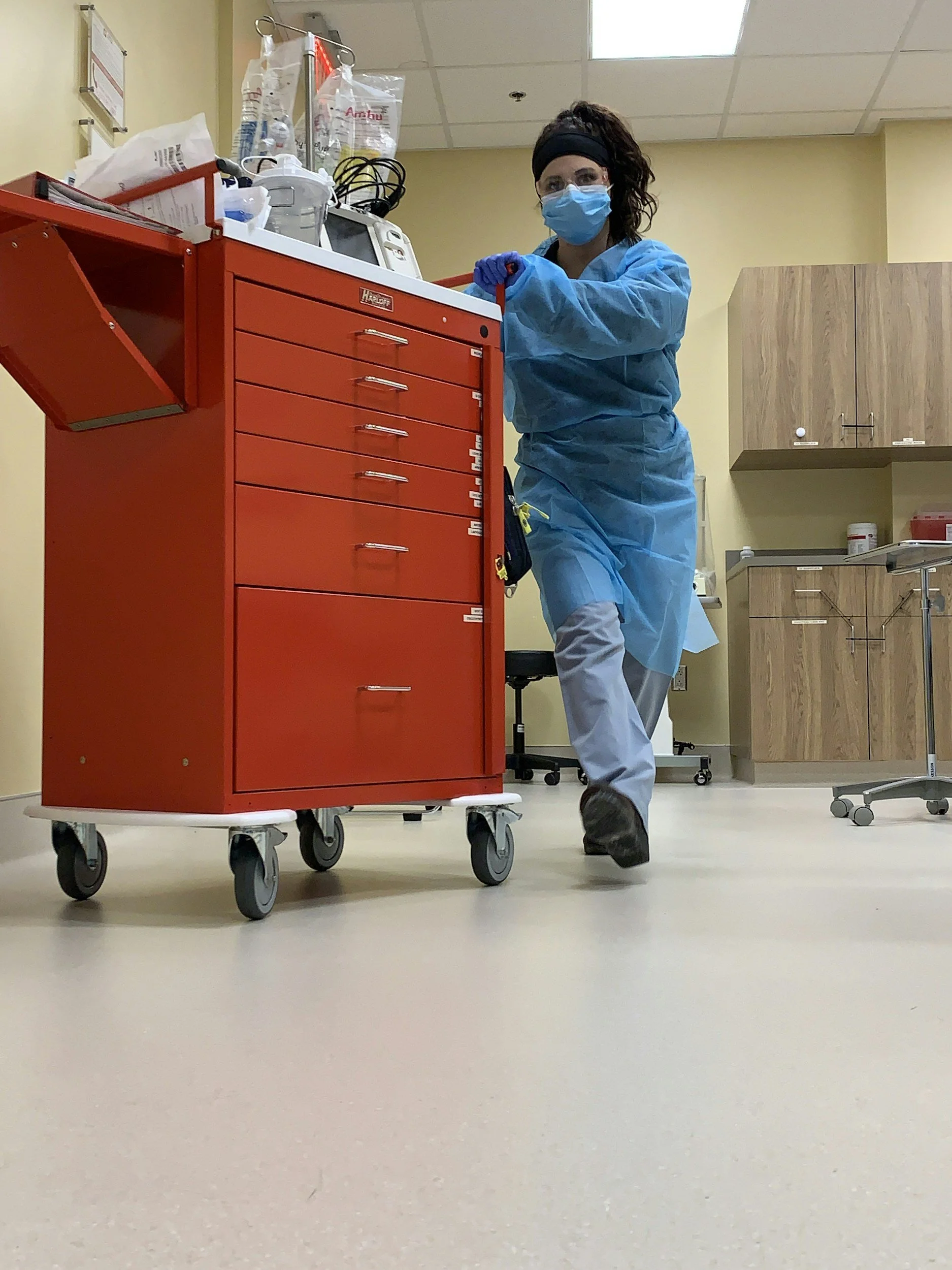 A healthcare worker in blue protective gear, face mask, and gloves stands next to a red medical cart in a hospital or clinic corridor, with cabinets and medical equipment in the background.