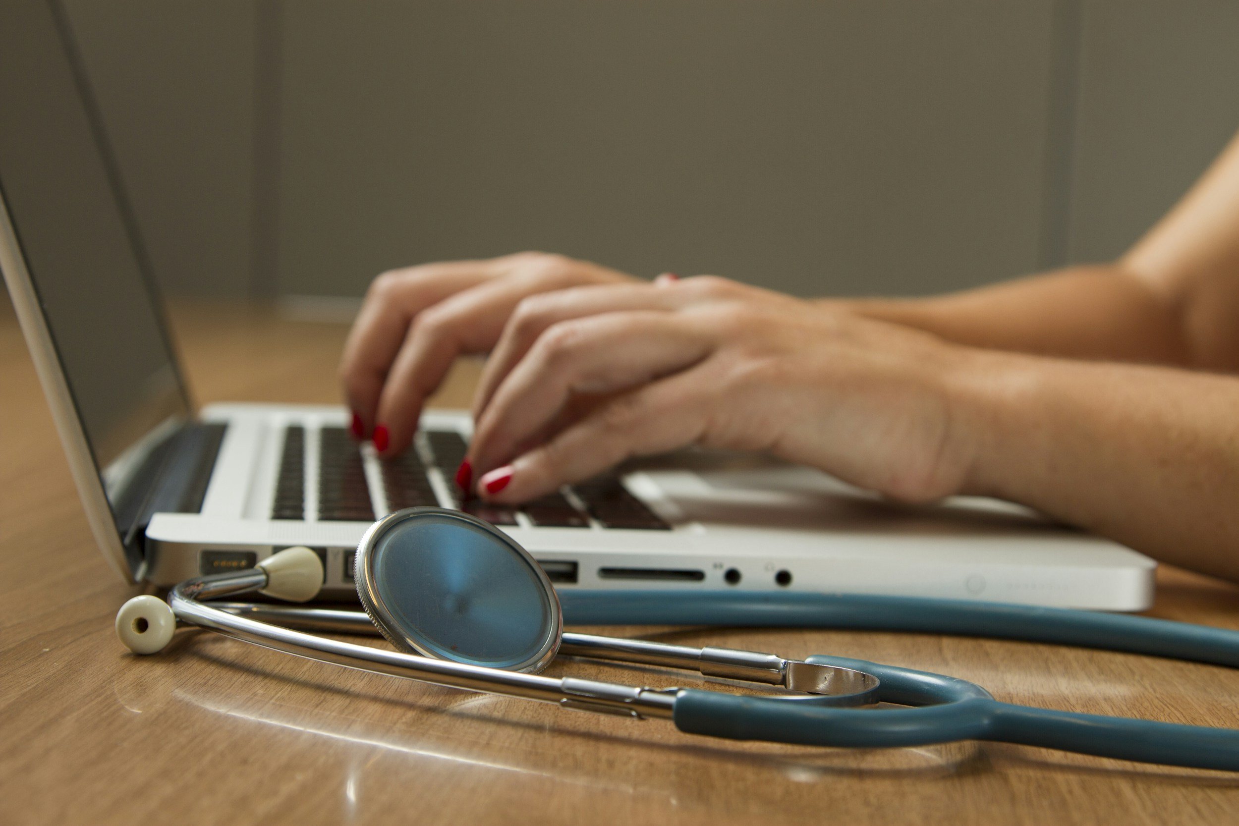 Close-up of hands typing on a laptop keyboard with medical stethoscope and earphones placed on a wooden table.