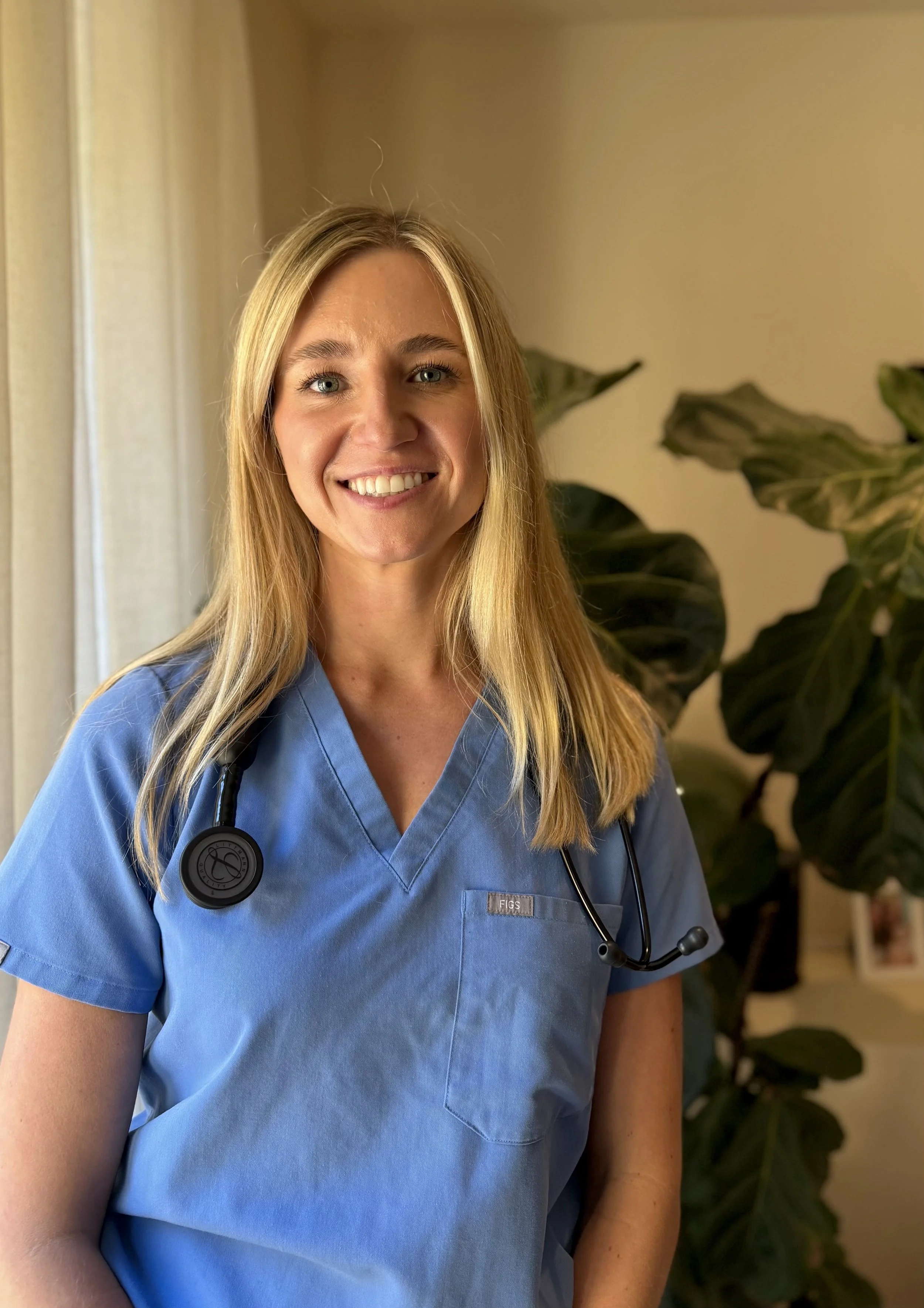 A smiling female healthcare professional wearing blue scrubs and a stethoscope around her neck, standing indoors with a plant in the background.