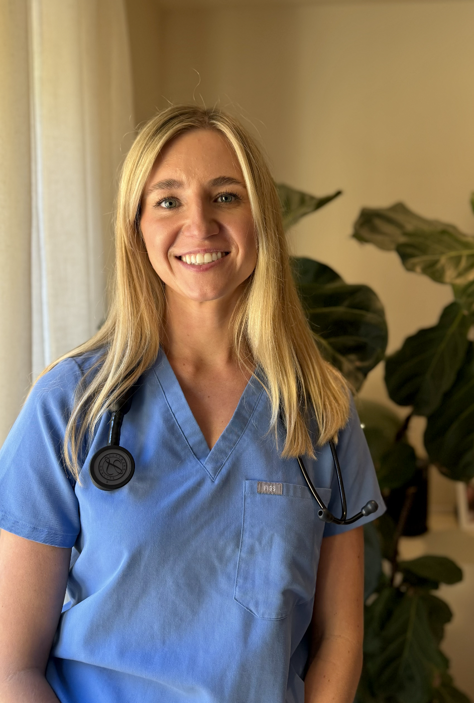 A smiling female healthcare professional in light blue scrubs with a stethoscope around her neck, standing indoors near a window and a large green plant.