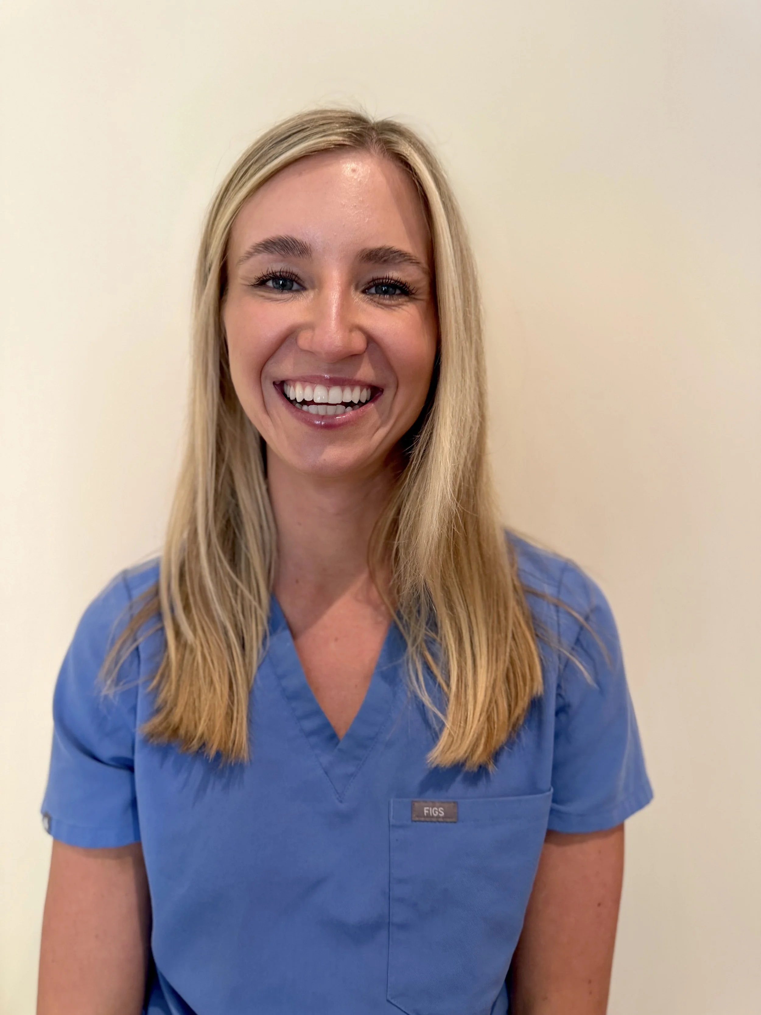A smiling blonde woman in blue medical scrubs standing against a plain off-white wall.