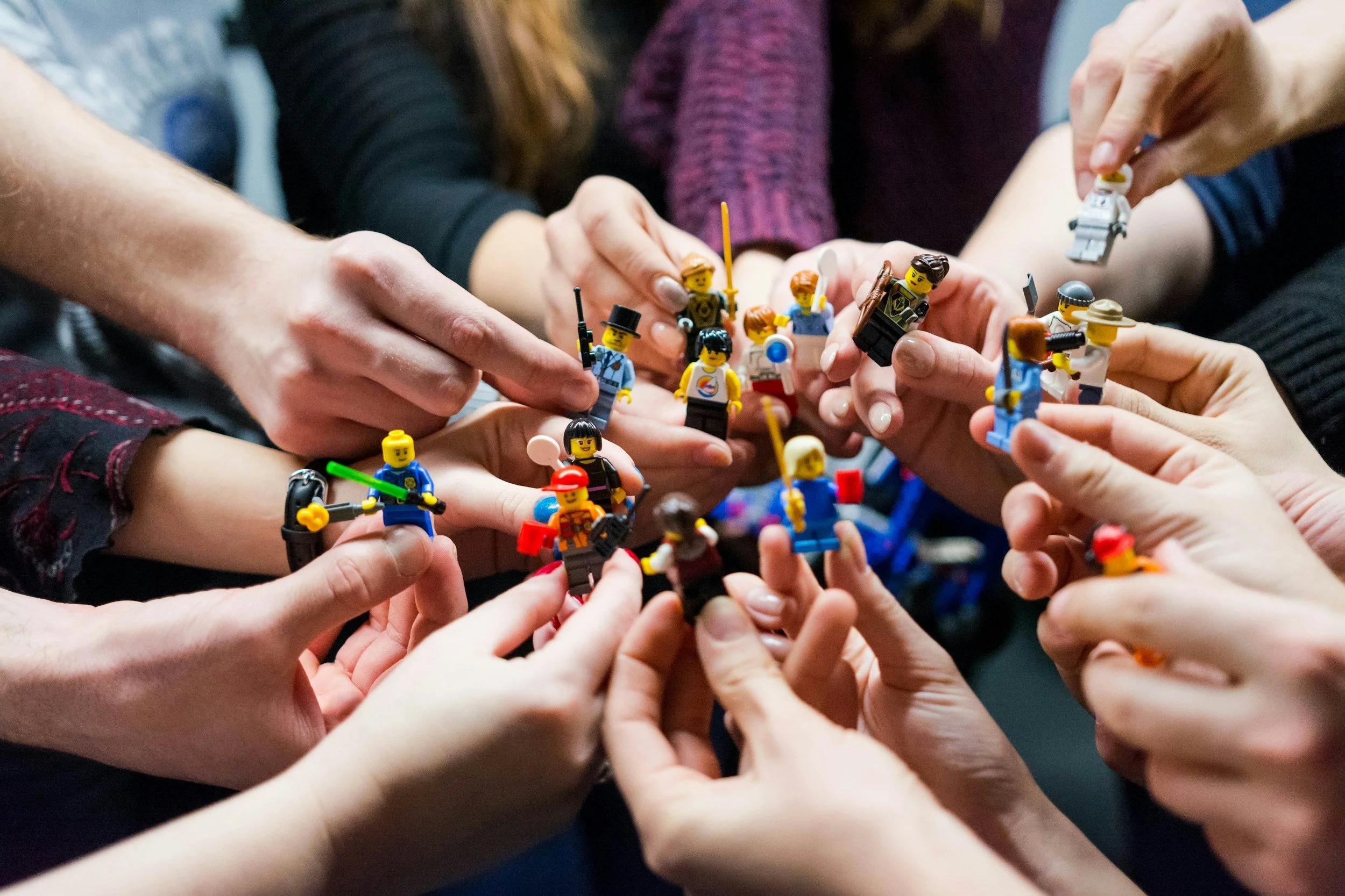 Multiple hands holding and arranging small LEGO figurines with varied costumes and accessories.
