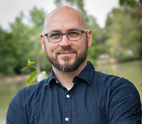 A man with a beard and glasses smiling outdoors, wearing a blue polka dot shirt with trees in the background.