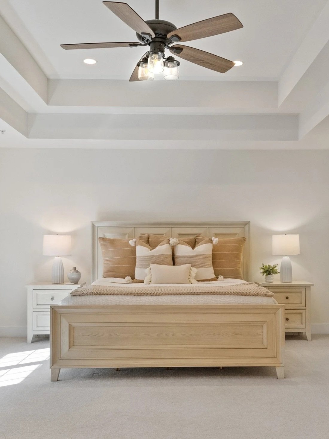 A bedroom with a light-colored wooden bed, beige pillows with pom-pom decorations, and white nightstands with table lamps, in a bright, neutral-toned room.
