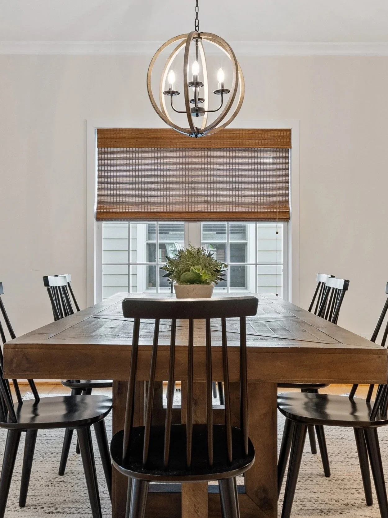 A dining room with a wooden table surrounded by six black chairs, a chandelier hanging above, a window with a venetian blind, and a potted plant on the table.