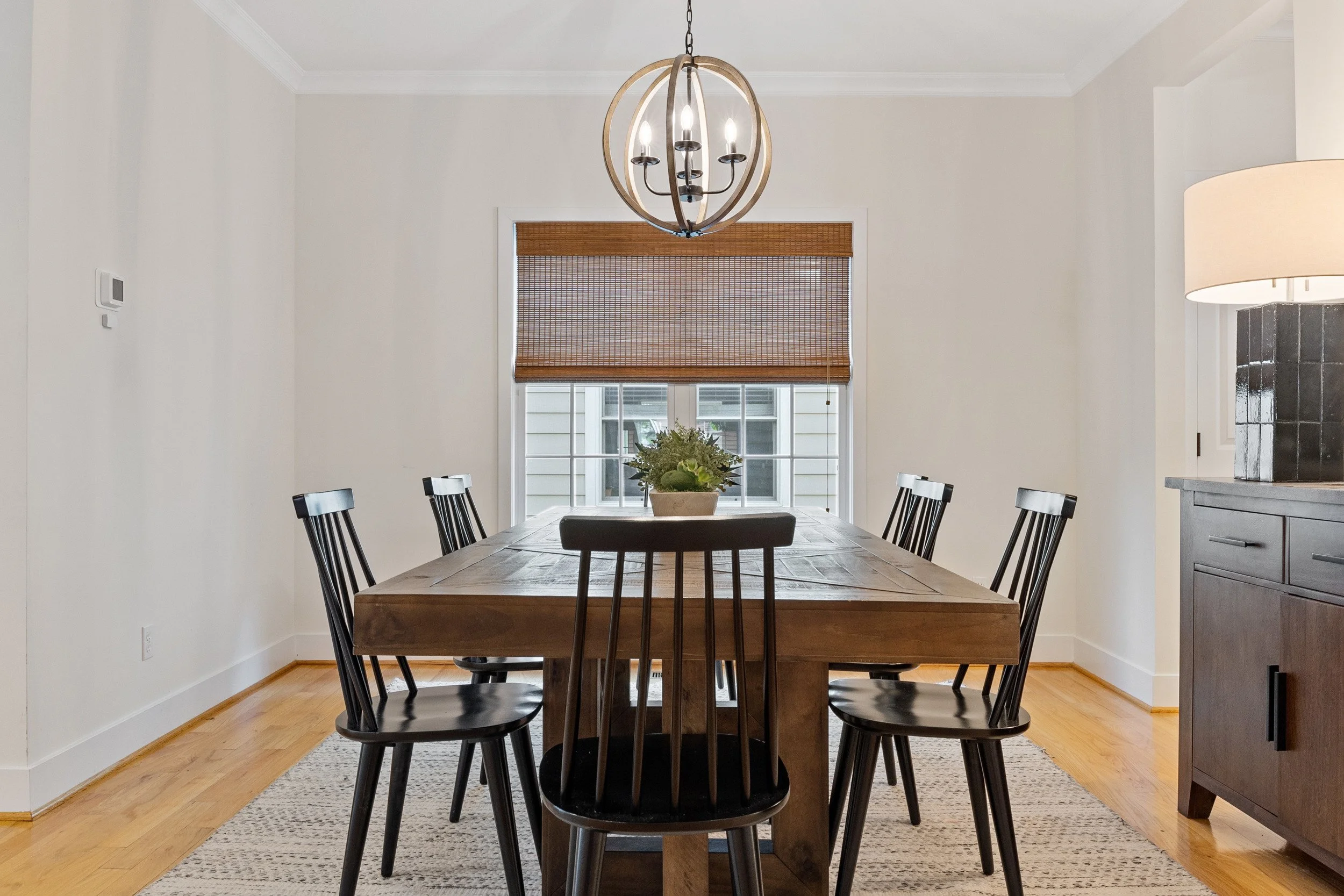 The Goat Getaway Dining room with hardwood flooring, a rectangular wooden table, black spindled chairs, a woven window shade, a chandelier, a potted plant, and a sideboard.