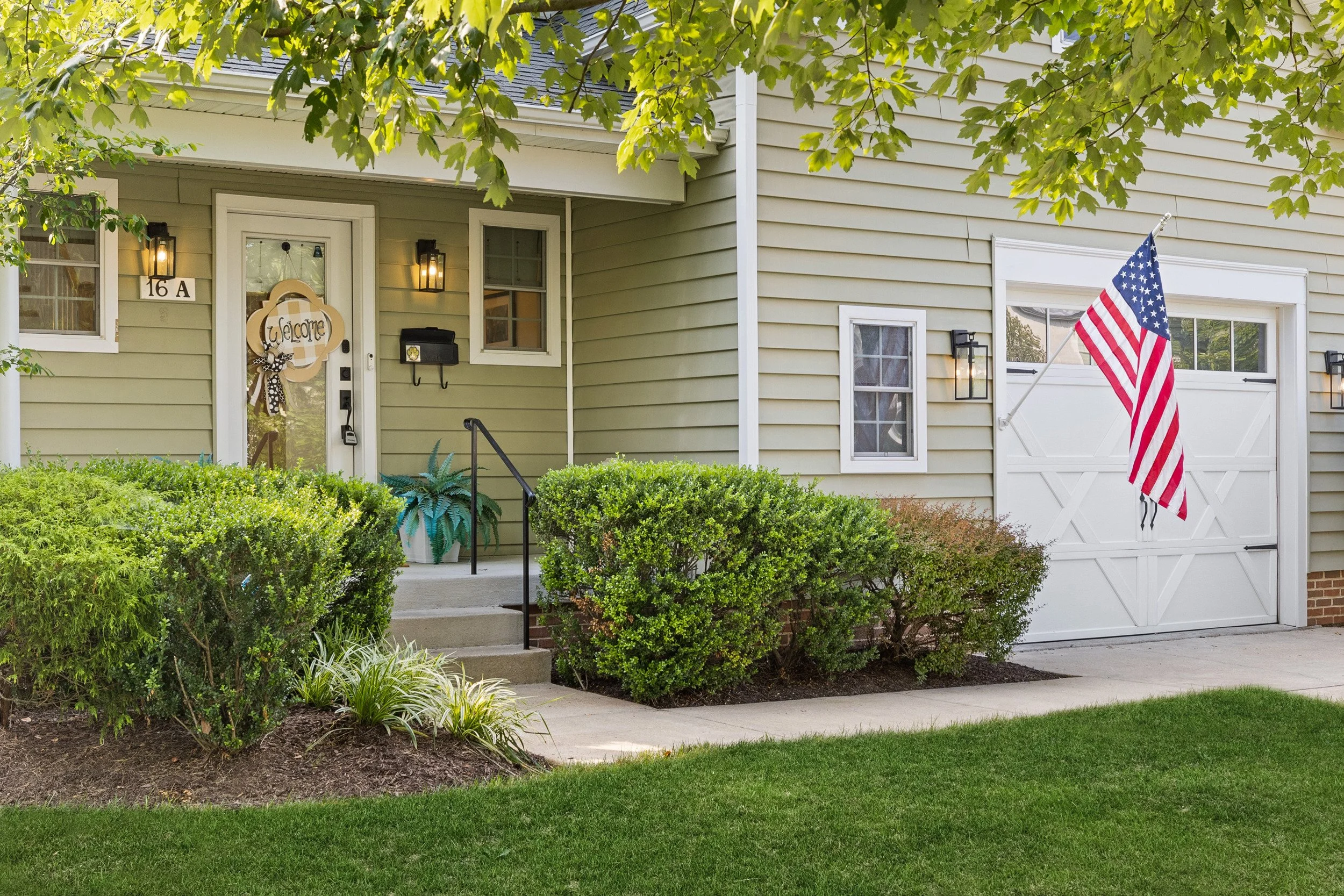 The Goat Getaway, a vacation rental's front exterior features a beige house and a garage door, an American flag, and lush green bushes and trees.