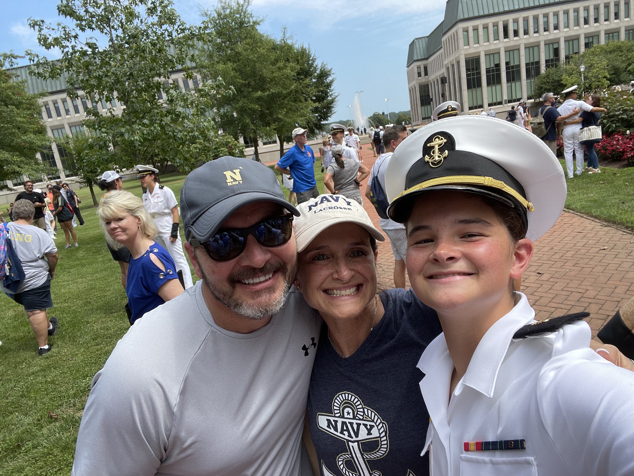 The Kelley family outdoors, one Navy uniforms, taking a selfie in front of a fountain and an office building.
