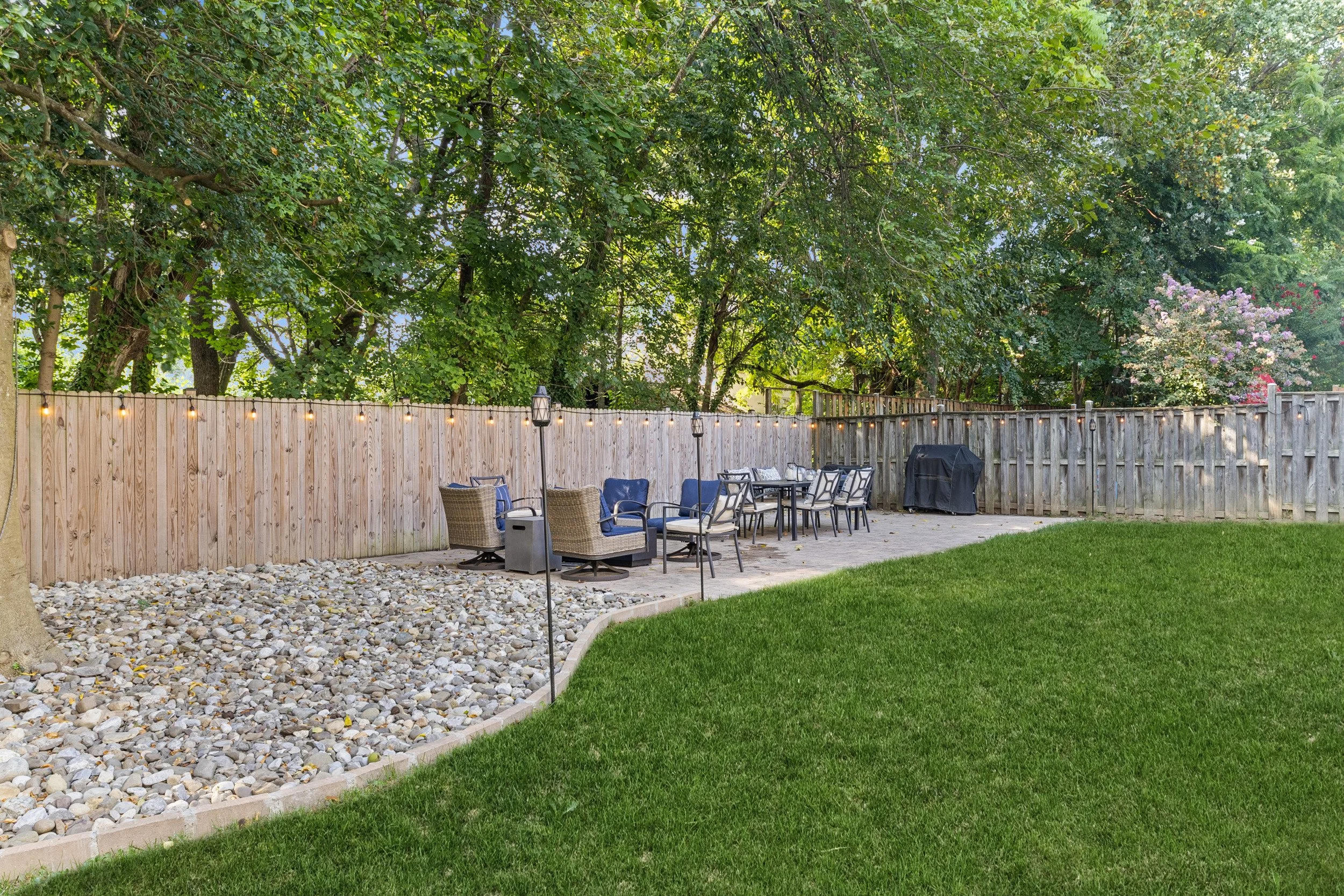 A backyard patio with outdoor furniture, a string light, and a wooden fence with lush green trees in the background.