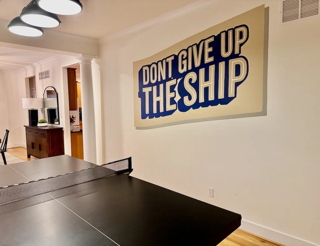 A wall in a home interior with a large sign that reads 'Don't give up the ship' in bold blue and white lettering. There is a ping pong table in the foreground and a dining area with a sideboard and decorative mirror in the background.