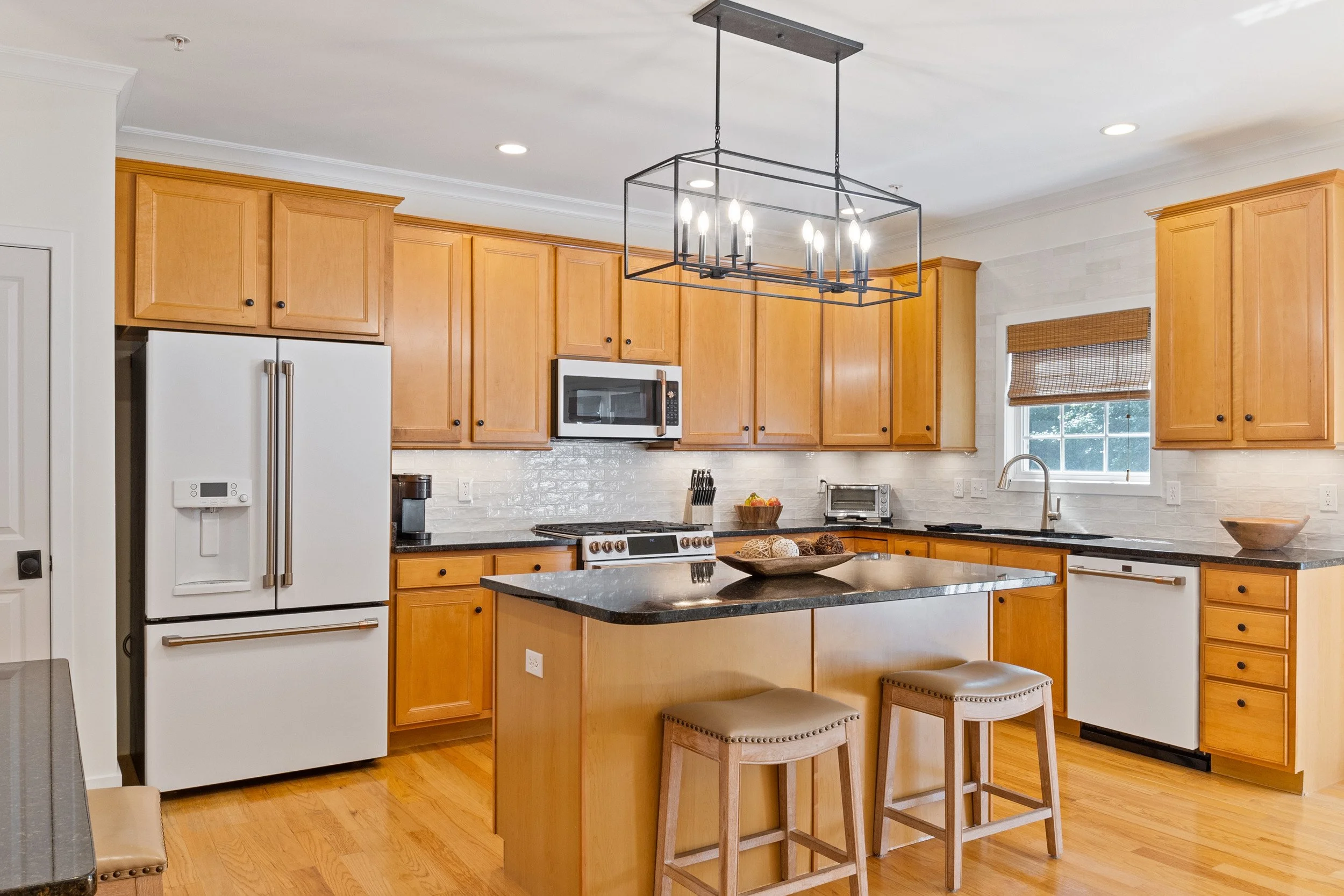 A bright kitchen with wooden cabinets, a white refrigerator, a built-in microwave, black countertops, and an island with seating. Overhead is a modern black chandelier with candle-like lights. There are two stools with beige cushions at the island, a