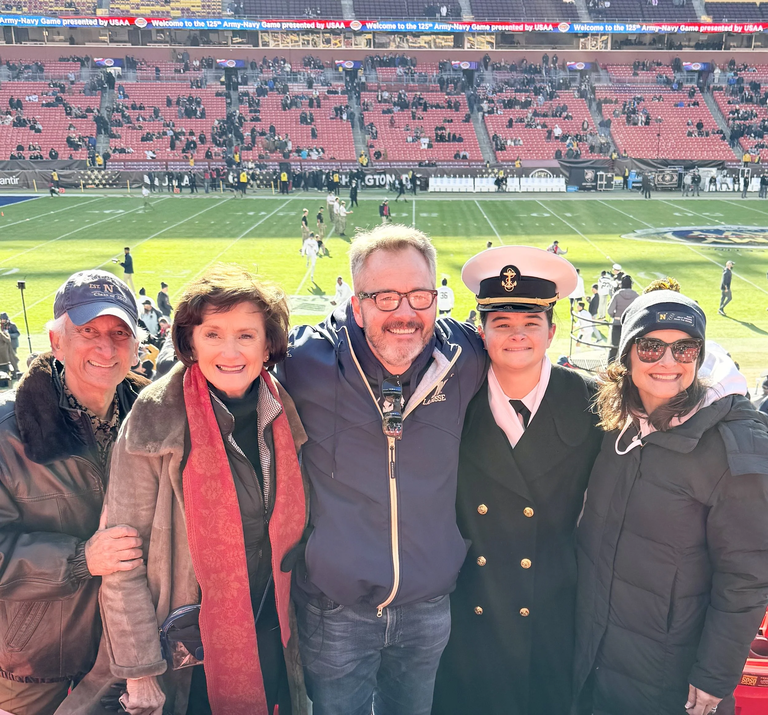 The Kelley family smiling at a football stadium, with some in military uniforms, during a game.