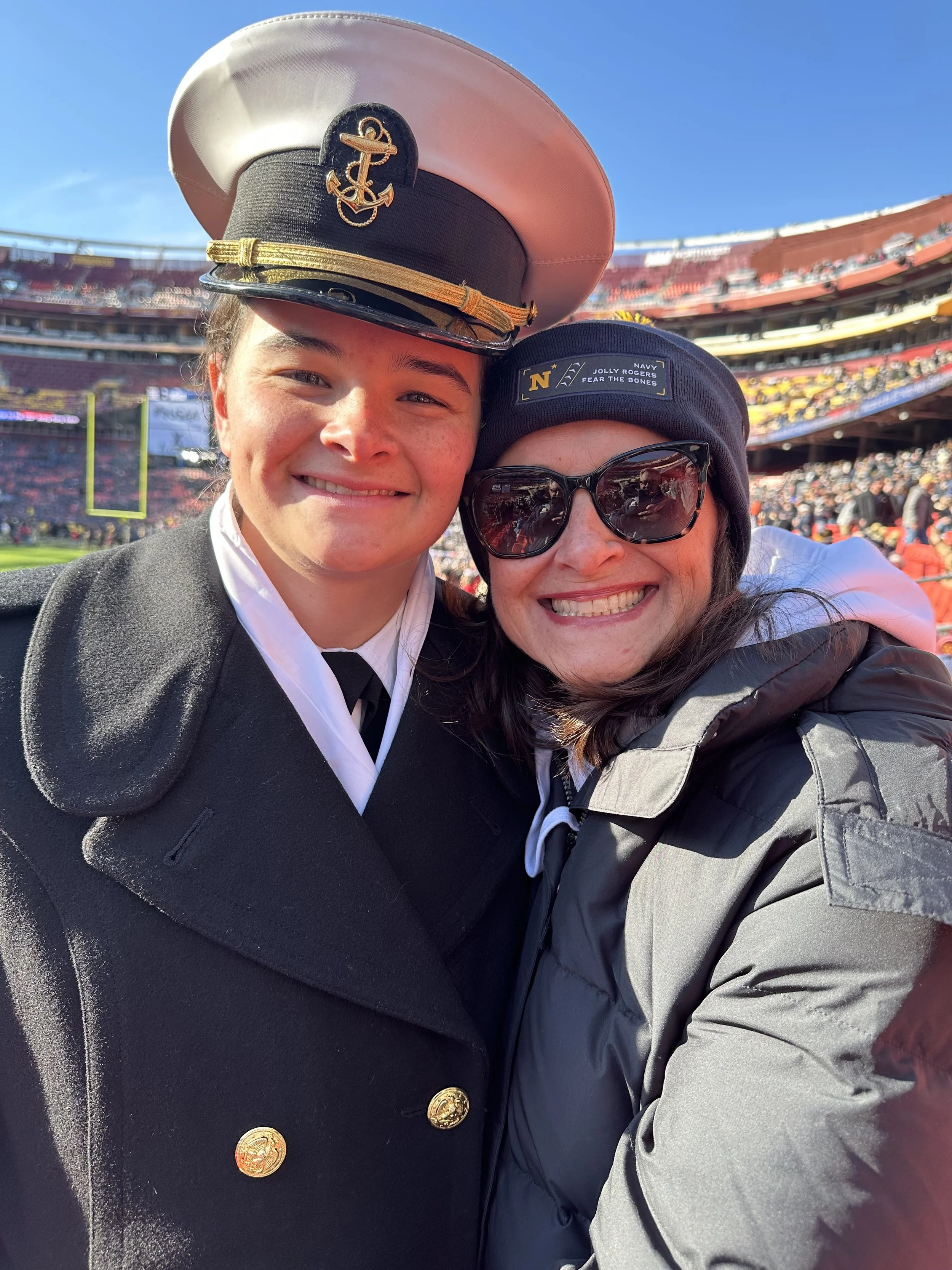 Kim Kelly and her daughter taking a selfie at a Navy Stadium, both smiling. Her midshipman is in uniform, and the stands are filled with fans on a beautiful day.