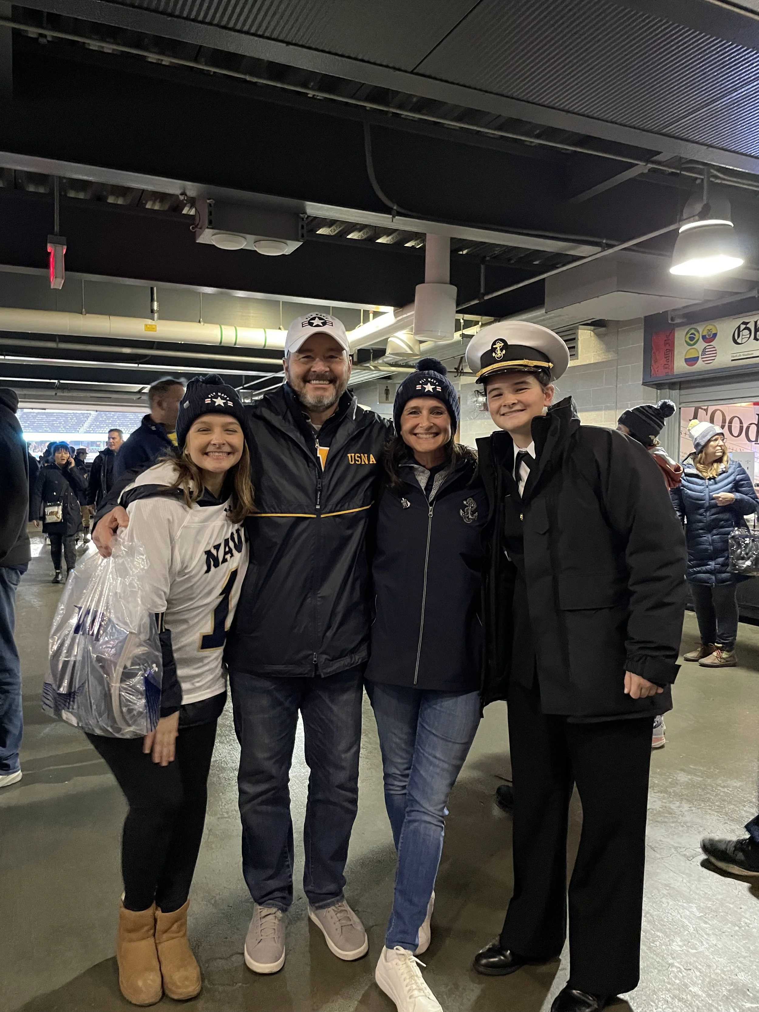 The Kelley family smiling and posing together inside Navy stadium. Two women, a man, and a young woman wearing a naval officer's uniform. The background includes other spectators and stadium lighting.