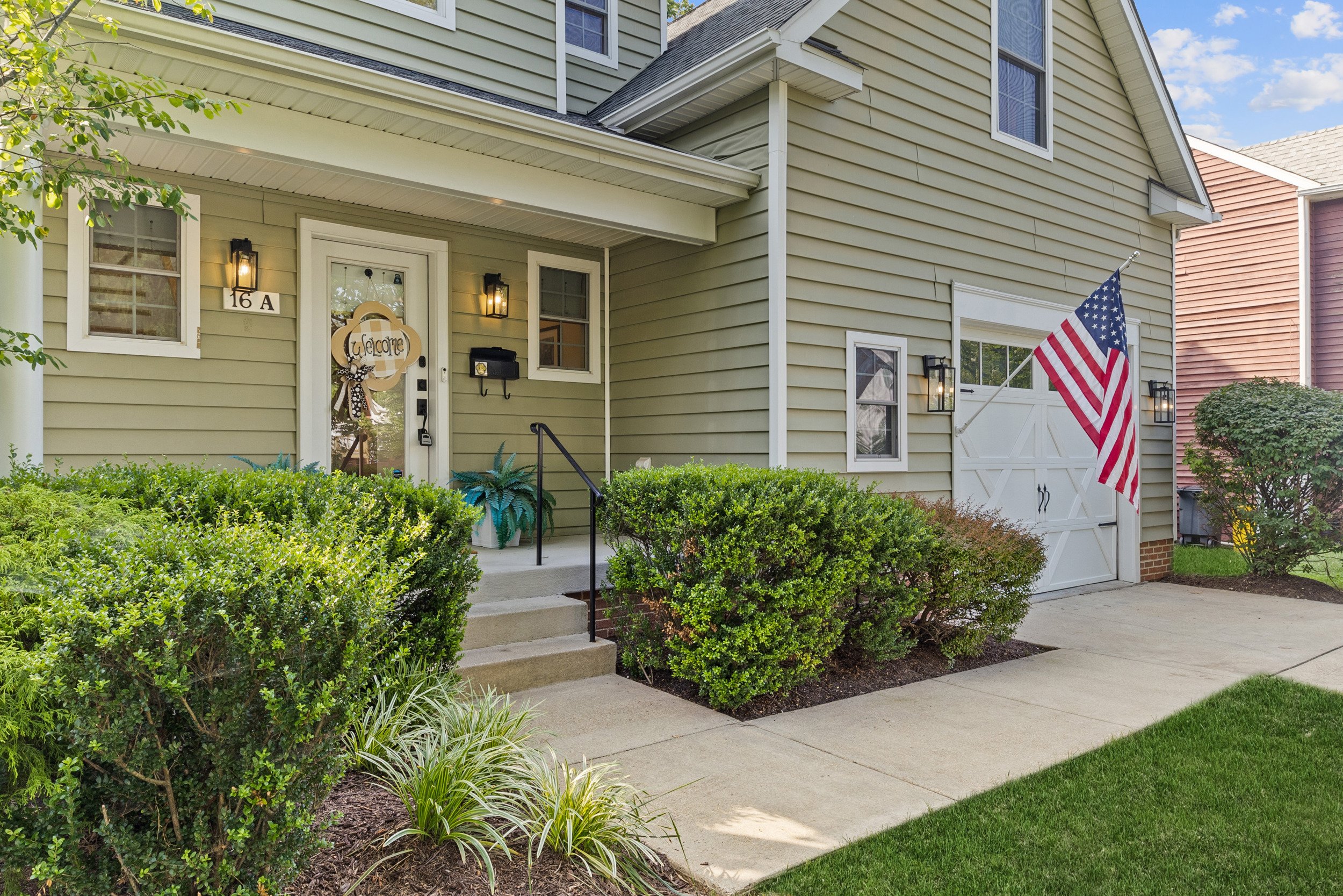 Front of a light green house with shrubs and plants in the front yard, American flag near the garage, welcome sign on the front door, and steps leading to the entrance.