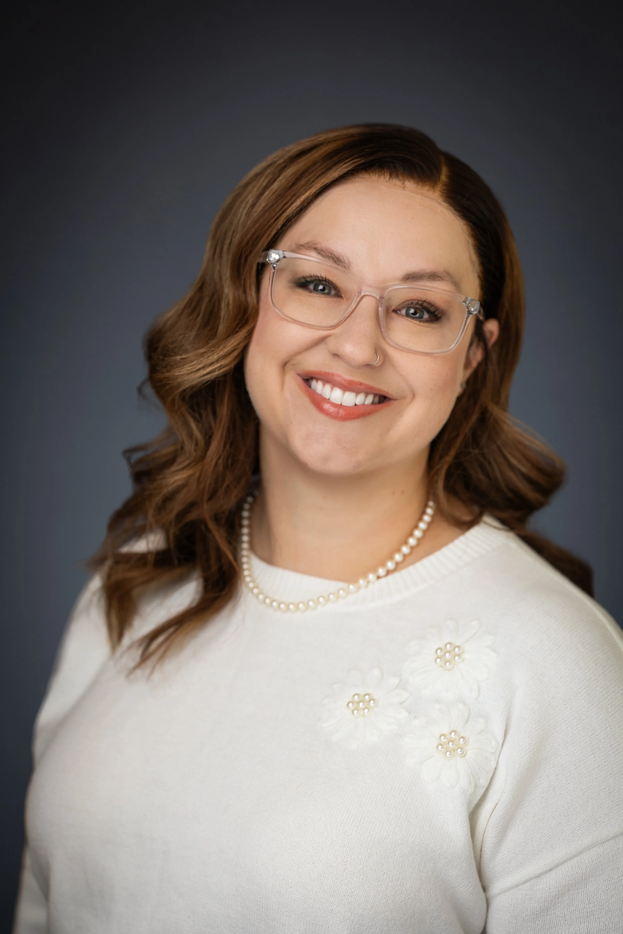 A woman with light skin, brown wavy hair, and glasses smiling, wearing a white sweater with floral embellishments and a pearl necklace, against a dark gradient background.
