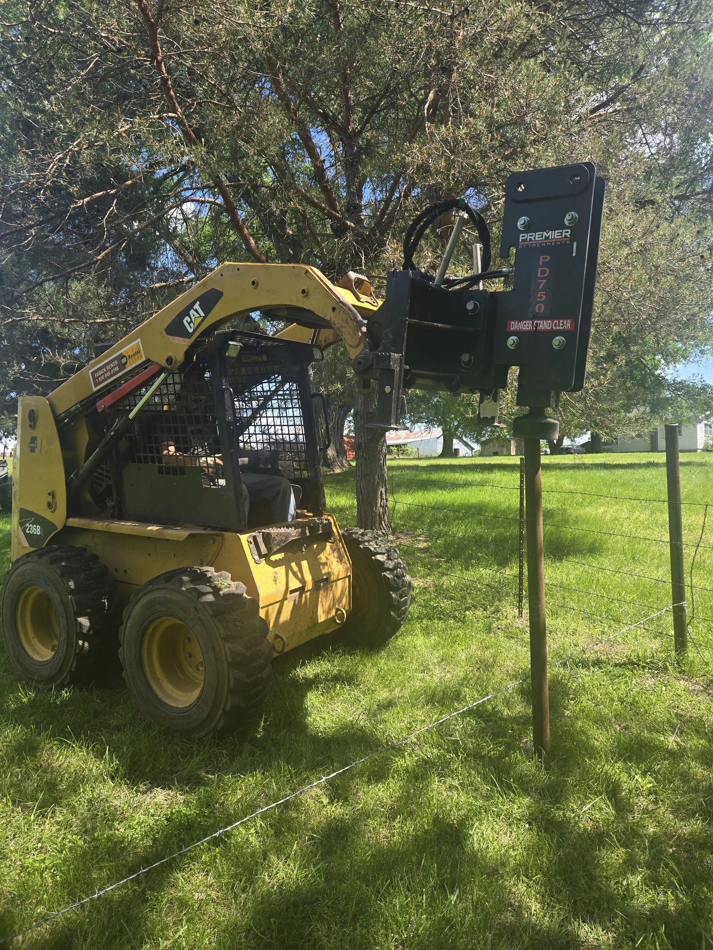 A yellow CAT construction vehicle skid steer is pounding in a pole in a grassy area with a tall tree and a fence nearby.