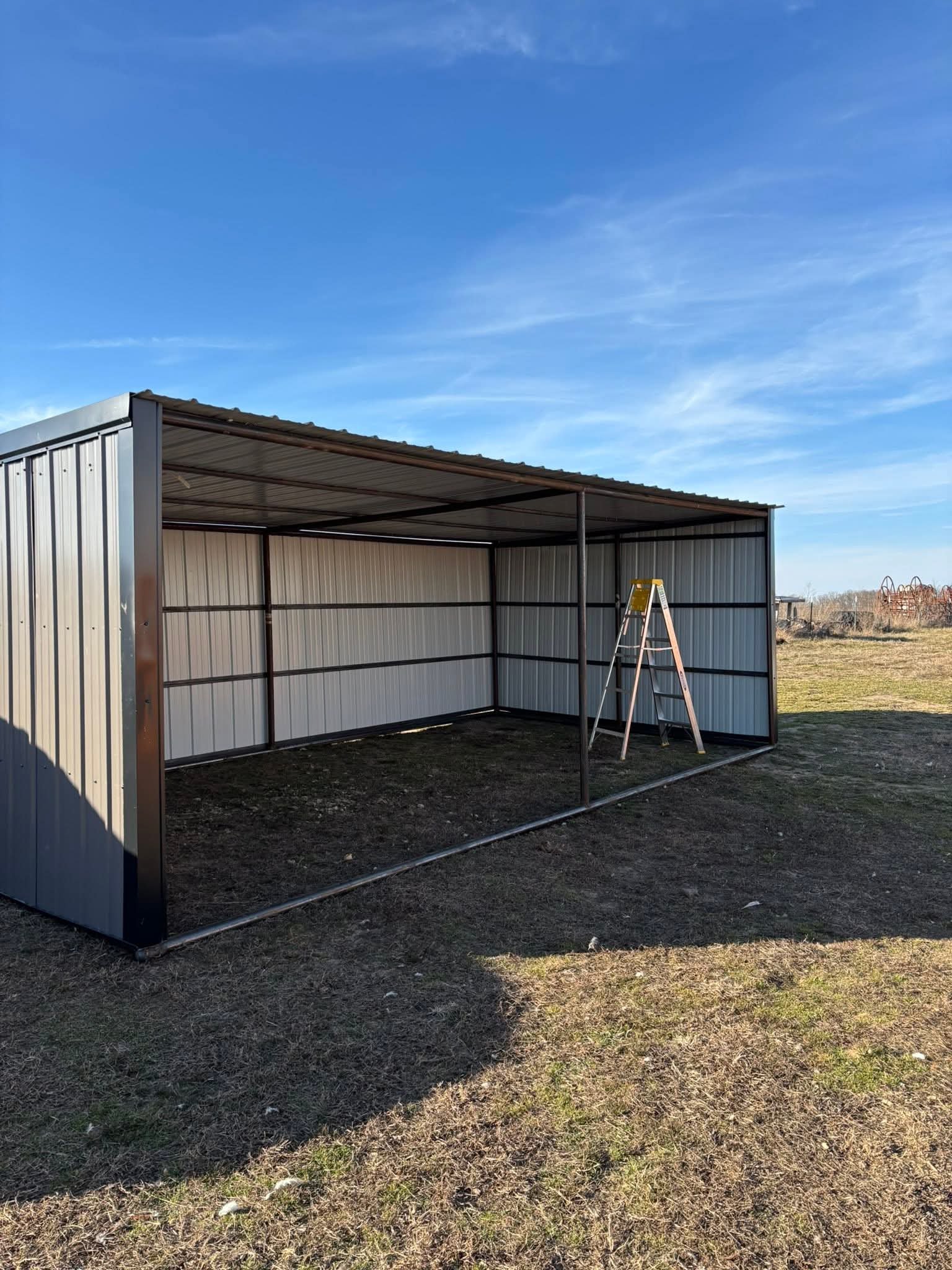 A metal animal shed frame with some of the siding attached, set on a grassy outdoor area under a blue sky with some clouds made for animals.