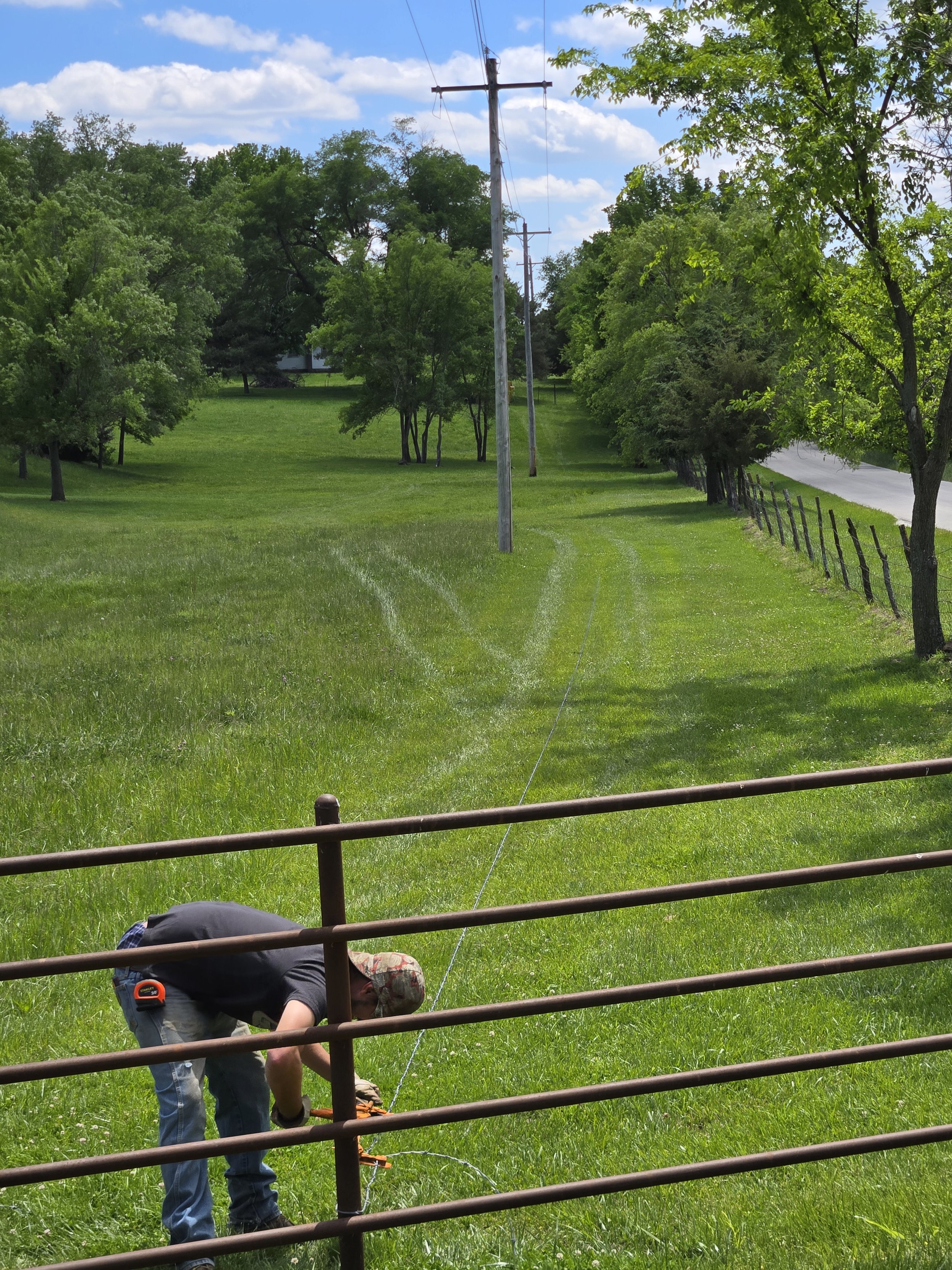 A person working with equipment in a grassy field next to a metal fence, with trees and power lines in the background under a partly cloudy sky.