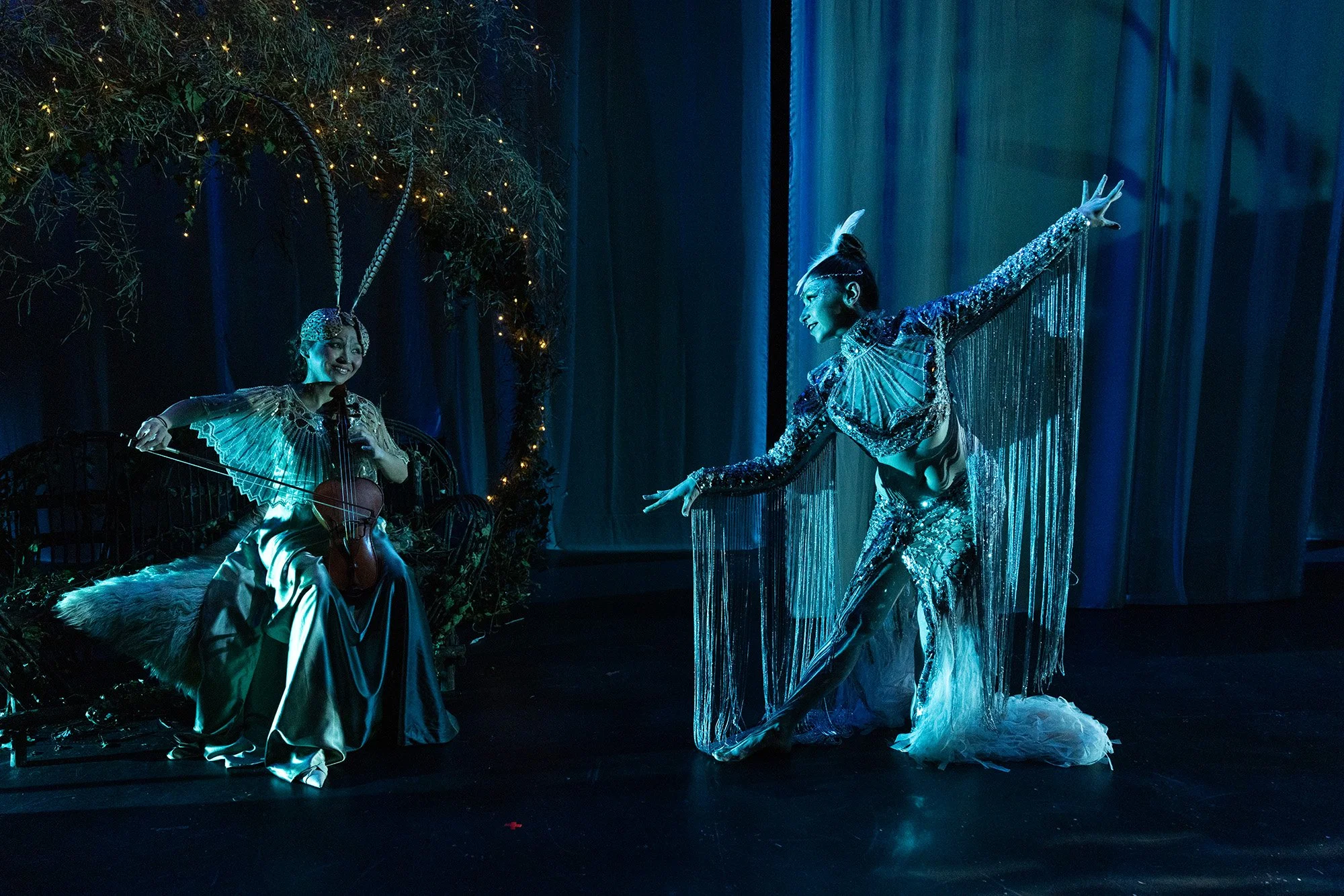 Two performers on stage, one seated playing a instrument and the other standing, both dressed in shiny, elaborate costumes with sparkling details. The stage is decorated with dark curtains, foliage, and fairy lights, creating a whimsical atmosphere.