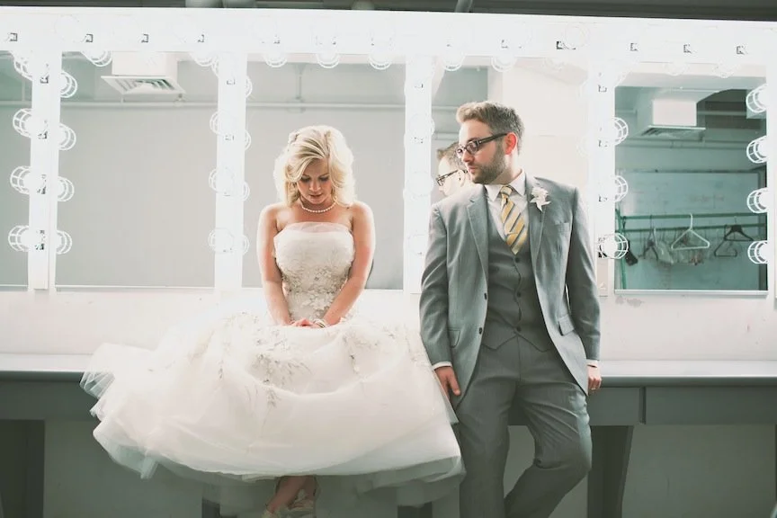 Bride in a white wedding gown standing next to a groom in a gray suit in a brightly lit dressing room.