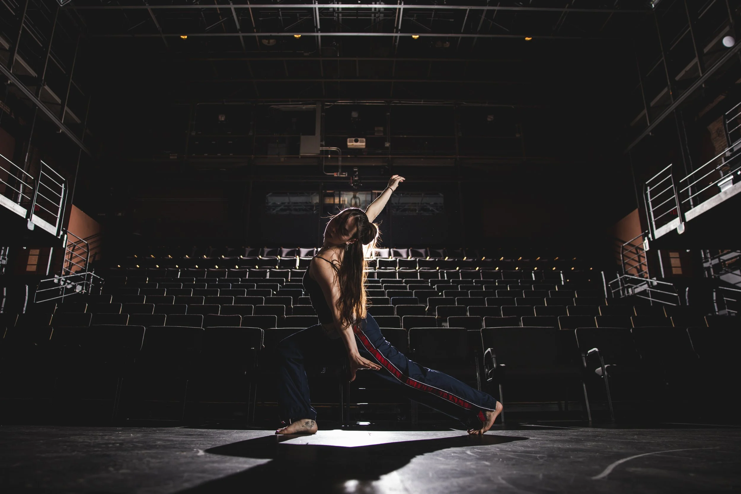 A woman practices dance routine on stage in an empty theater with rows of seats, dim lighting, and a black ceiling.