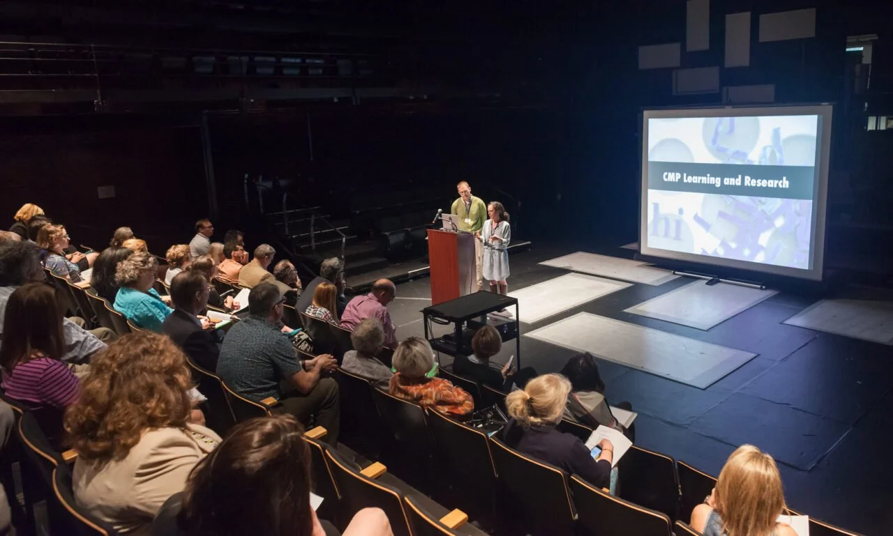 A corporate presentation with two presenters standing at a podium on the New Hazlett Theater stage in front of a large audience. A screen displays 'CMR Learning and Research' in the background.