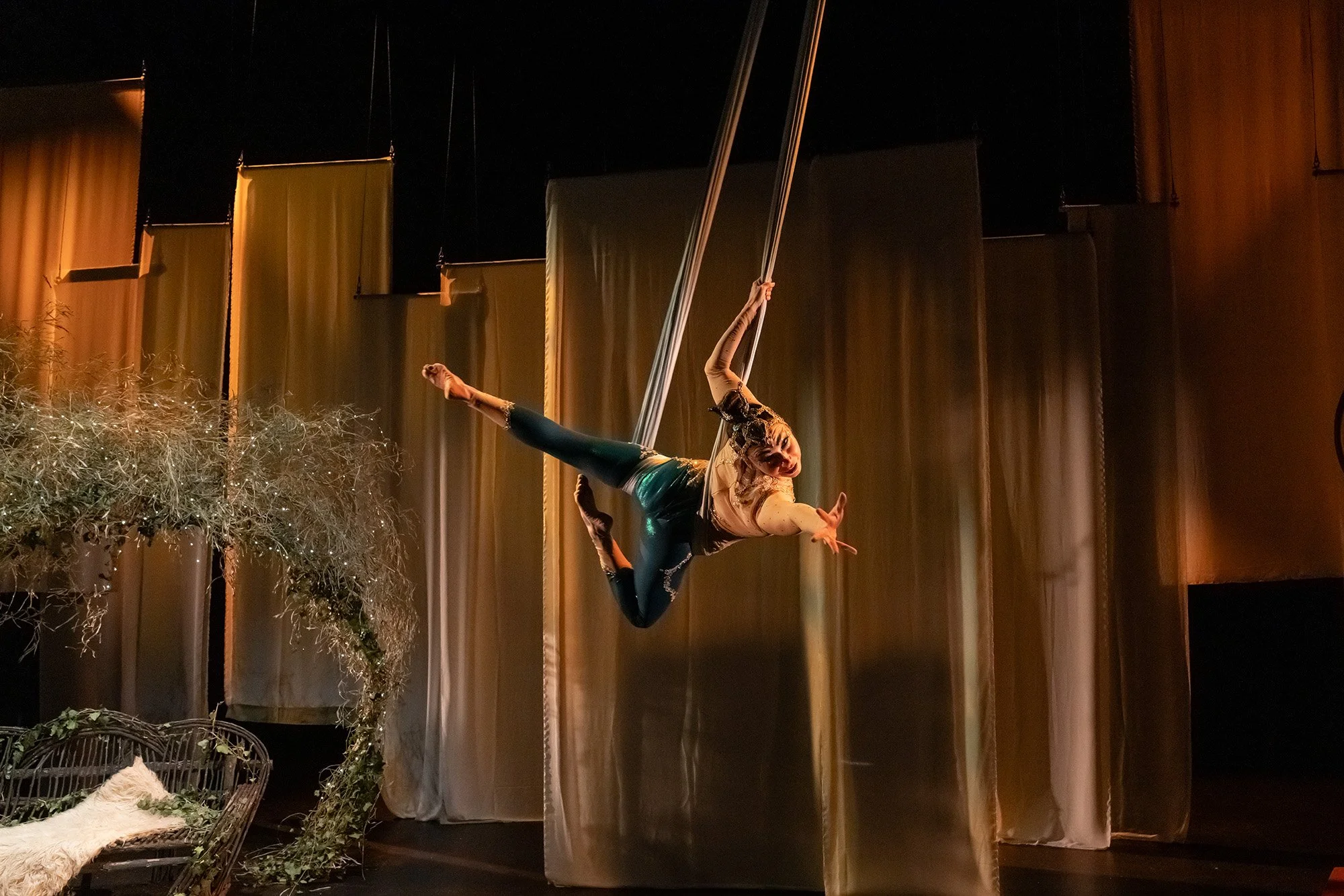 A woman performing an aerial silk routine on stage, hanging upside down with one leg extended and looking down, against a backdrop of curtains and stage decorations.