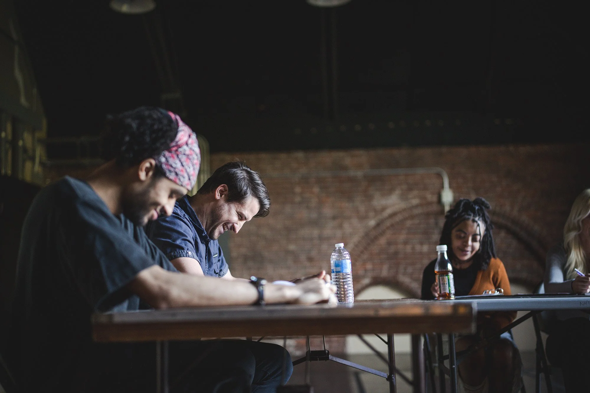 A diverse group of young adults sitting at a conference table, smiling and engaging in a discussion, with water bottles and soda on the table, in an industrial-style room with brick walls.