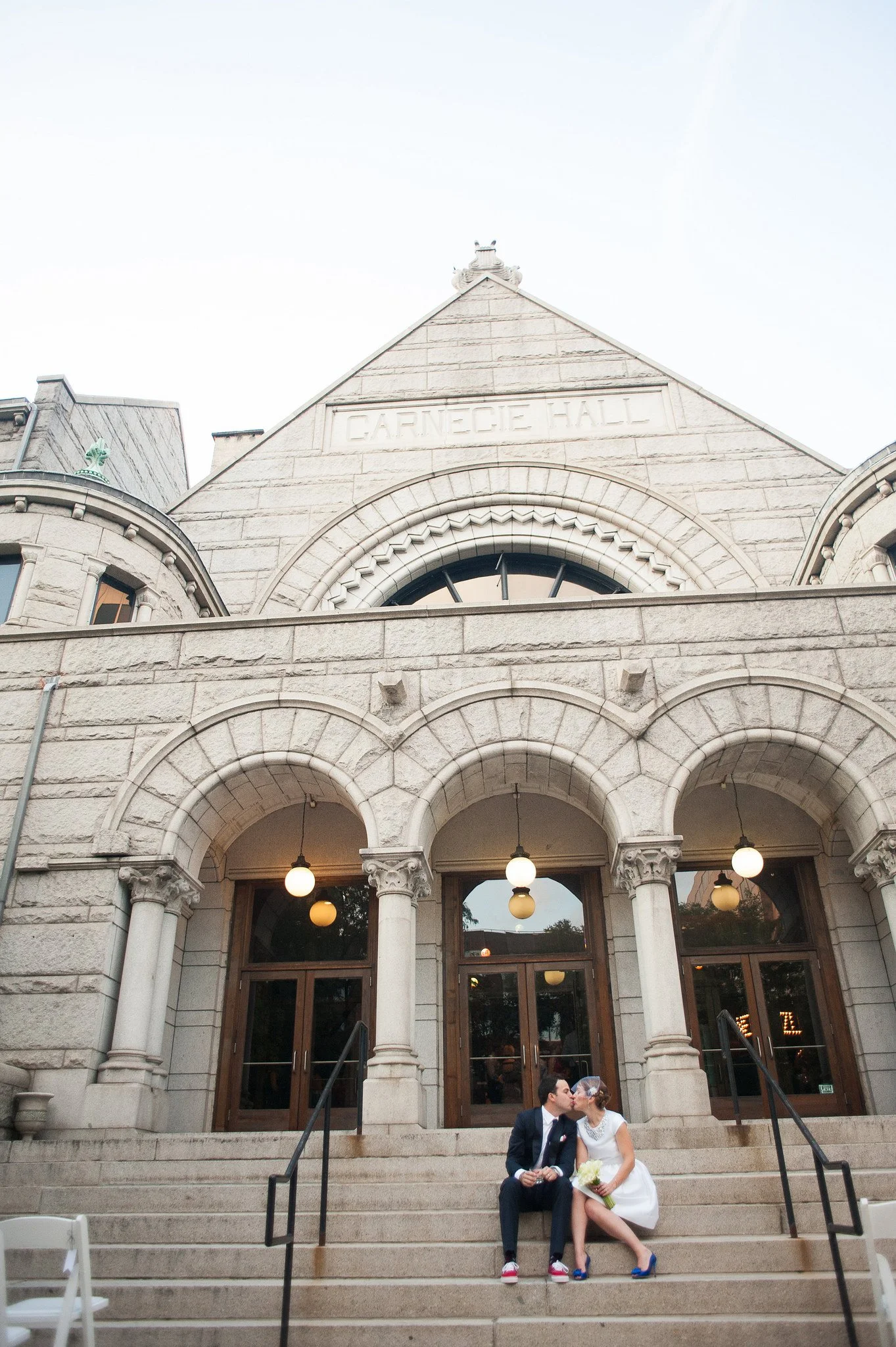 A couple dressed in wedding attire sitting on steps outside a historic stone building named Carnegie Hall, sharing a kiss. The bride holds a bouquet and wears a white dress and veil, while the groom is in a dark suit and sneakers.