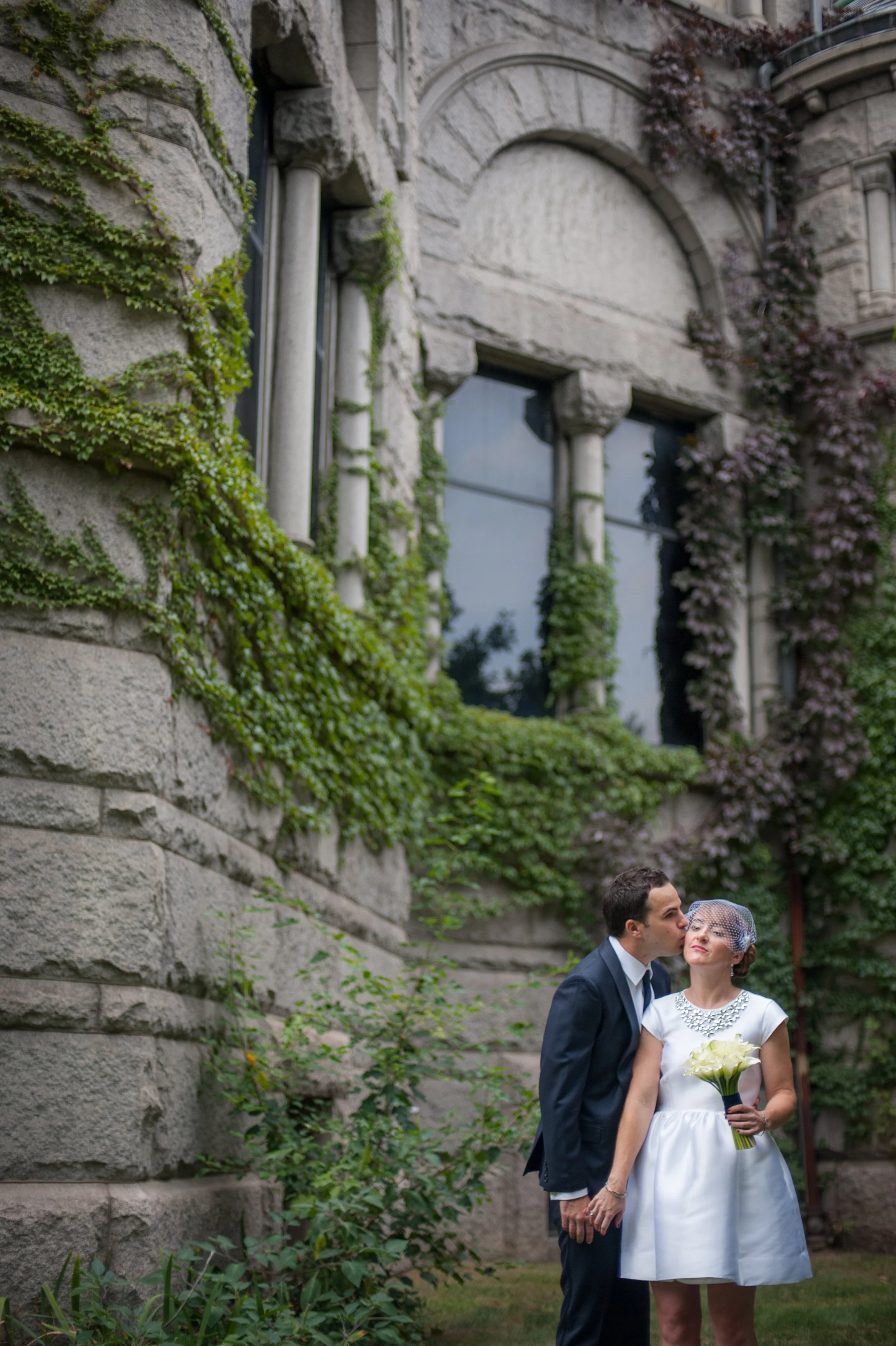 A newlywed couple standing outdoors in front of a stone building with ivy. The groom, in a dark suit, kisses the bride, who is in a white dress holding a bouquet of yellow roses, on the cheek.