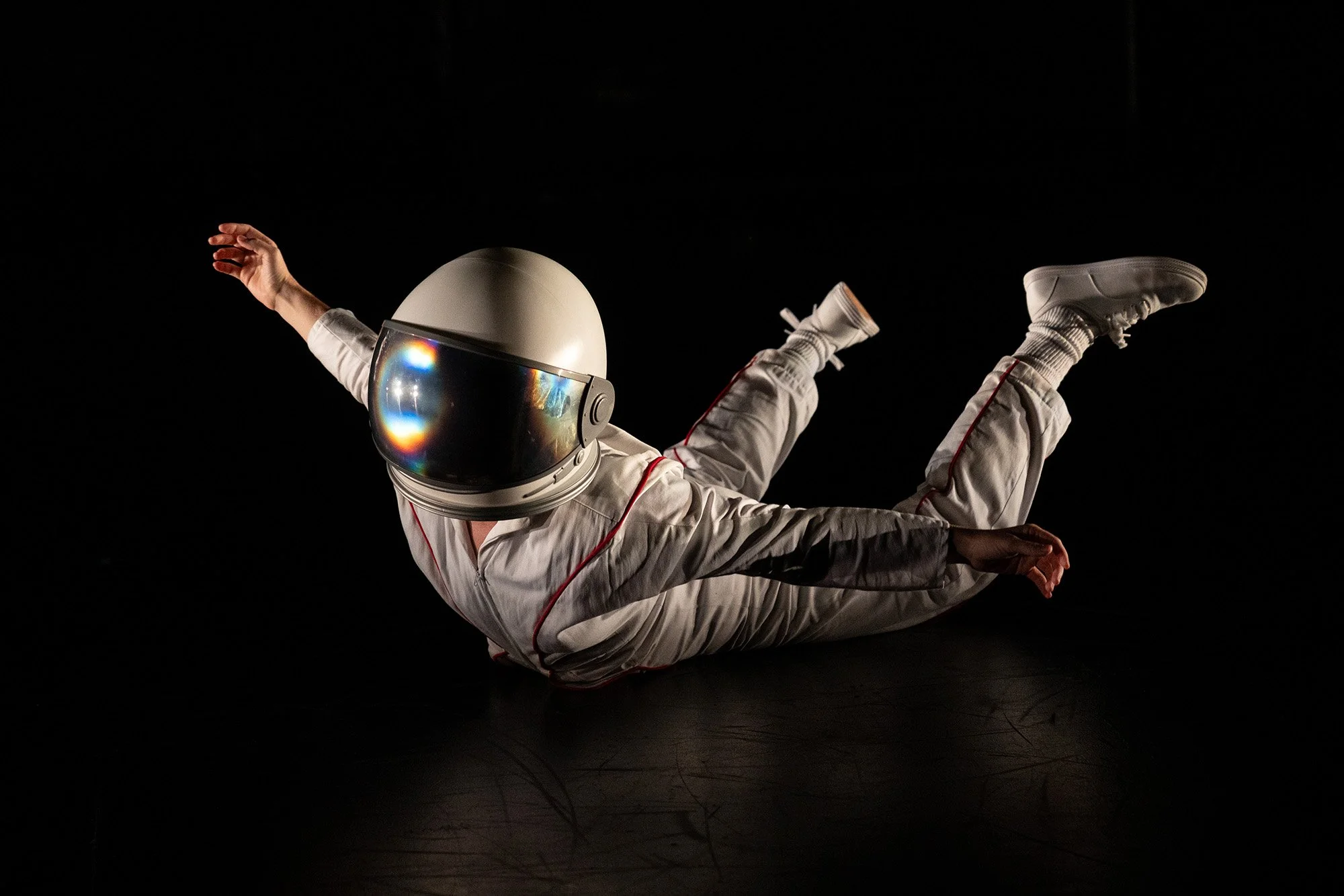 Person in a white spacesuit and helmet performing a dance move in a dark space with a black background.