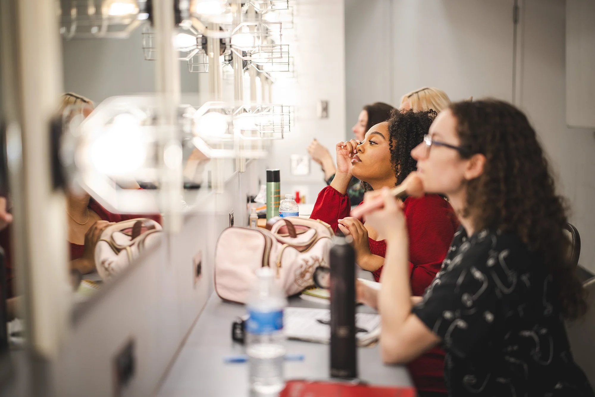 Women sitting at a makeup mirror with lights in the New Hazlett Theater doing their makeup with various brushes and tools.