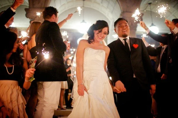 Wedding celebration with a couple holding hands and smiling, surrounded by guests holding sparklers on the steps of the New Hazlett Theater