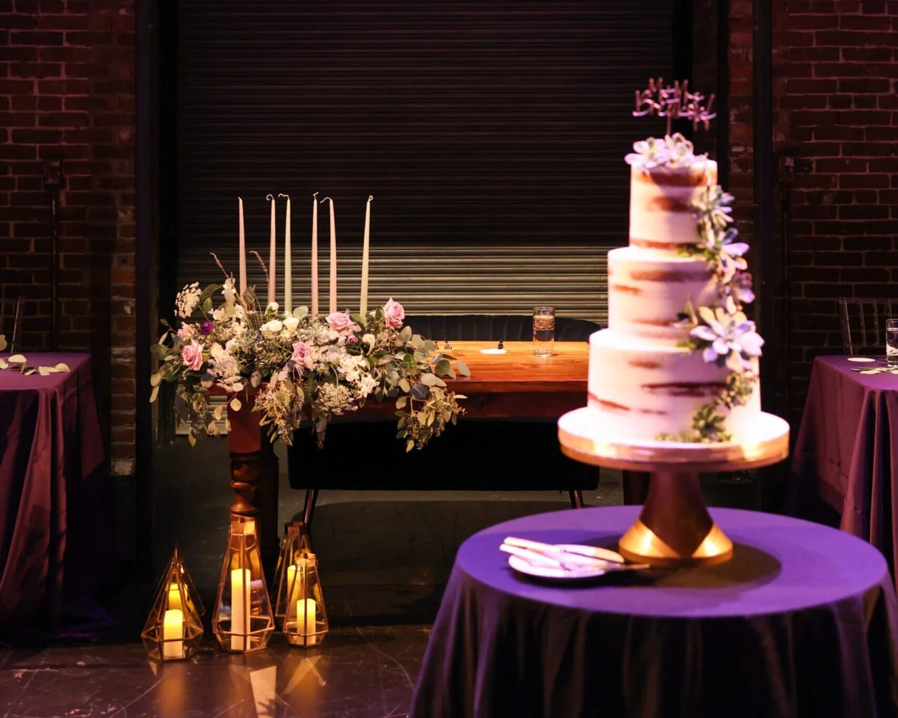 A wedding cake with three tiers decorated with flowers on a round table, with a floral arrangement and candles on the table behind it, in a decorated event space with purple drapes and brick walls.