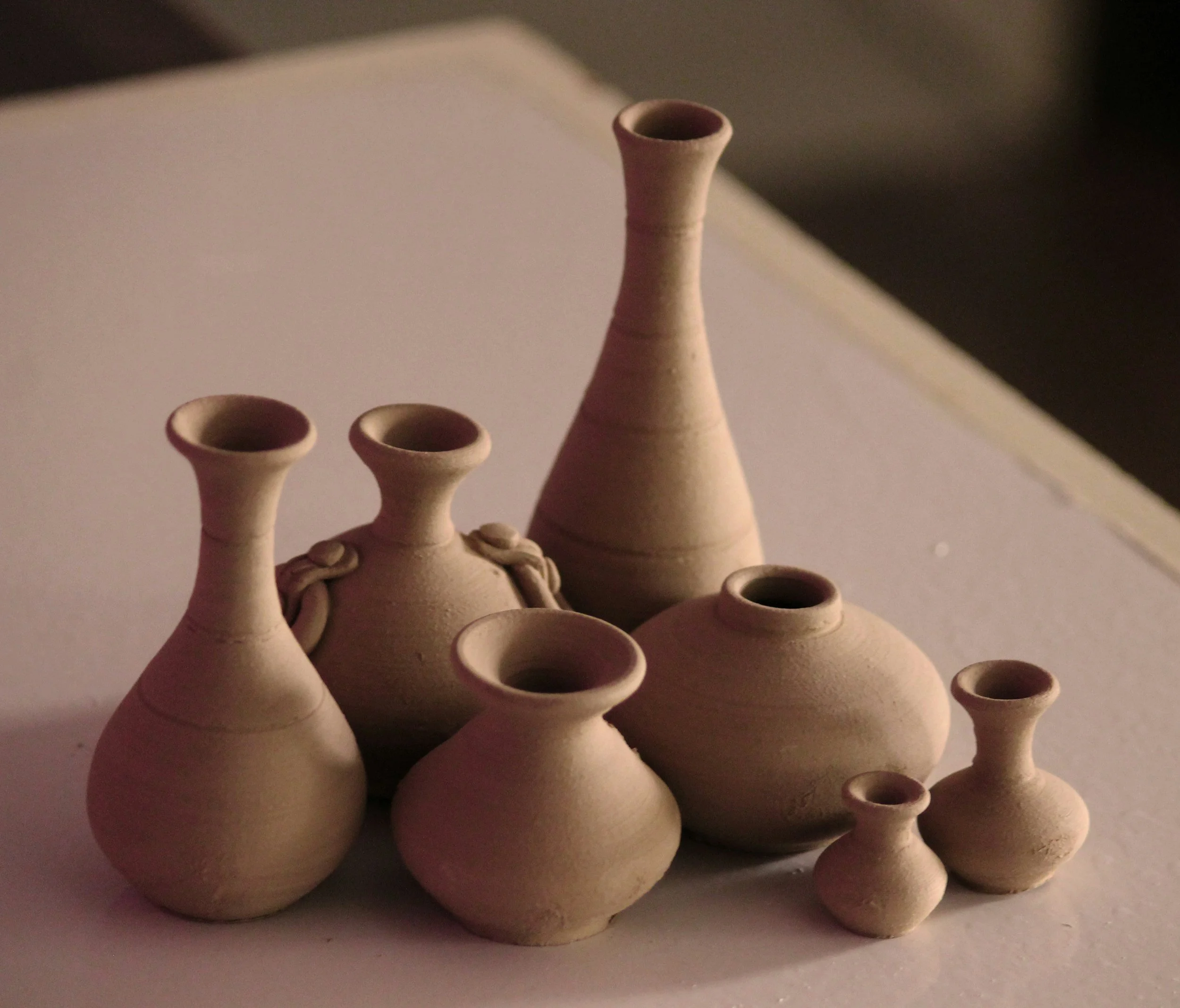 Multiple small ceramic vases and bottles in various sizes, beige color, arranged together on a flat surface, with a neutral background.
