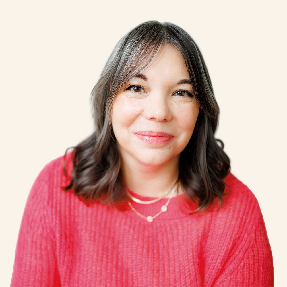 A woman with shoulder-length dark brown hair and a fair complexion, wearing a red sweater and a delicate necklace, smiling softly at the camera against a plain light background.