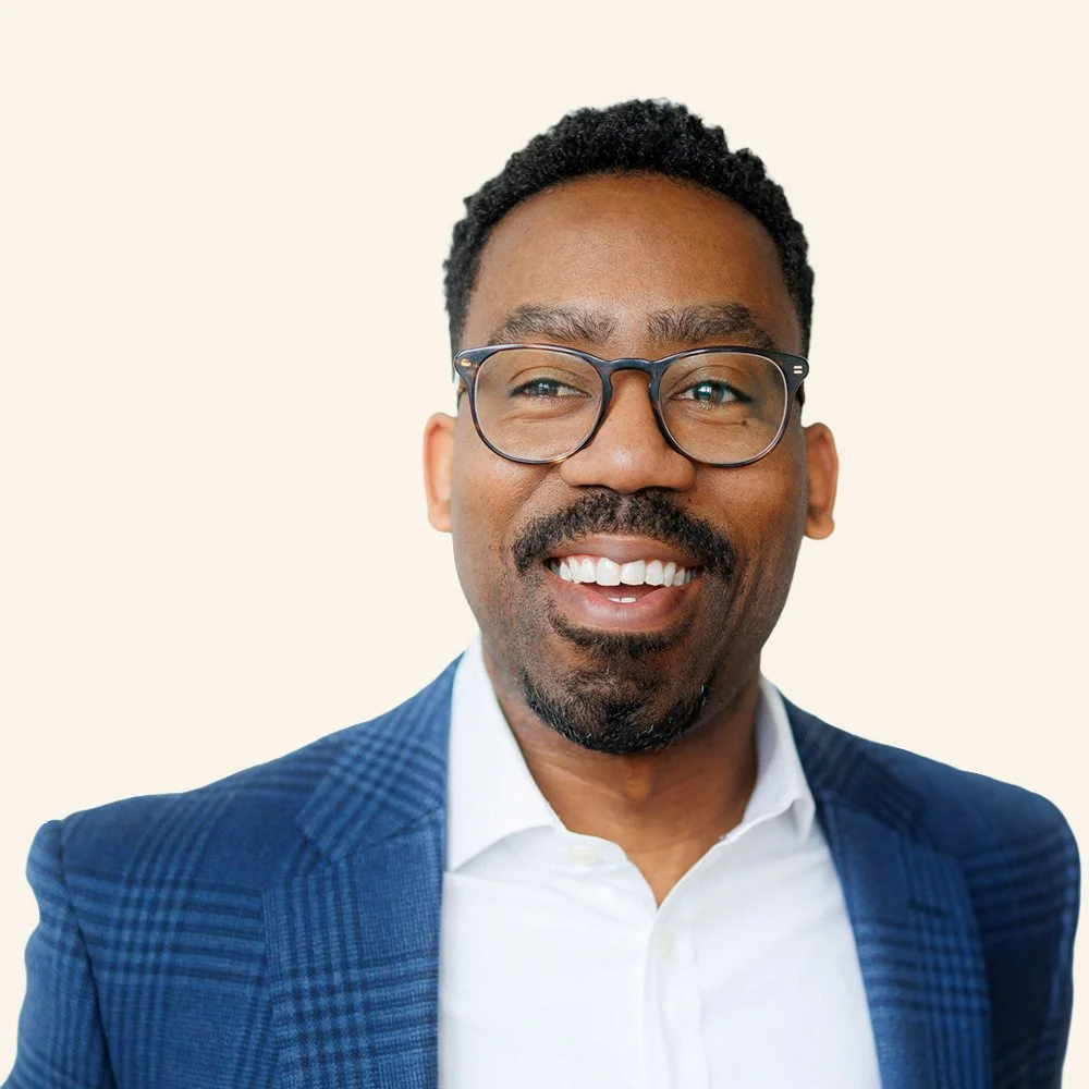 Smiling African American man with glasses, wearing a checkered blue blazer and white shirt against a plain background.