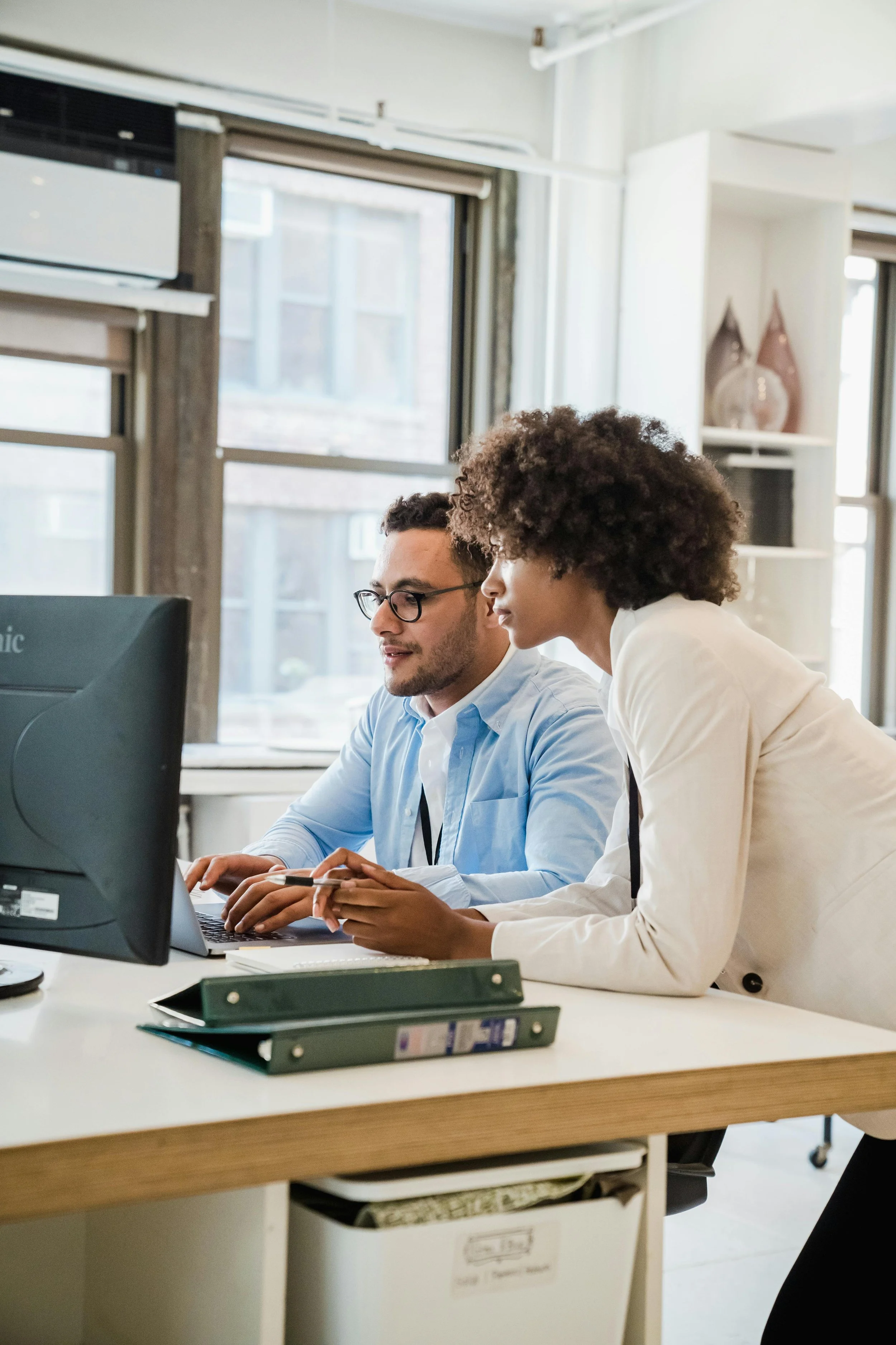 Two office workers, a man with glasses and a woman with curly hair, are working together at a desk with a computer in a modern office space with large windows and natural light.