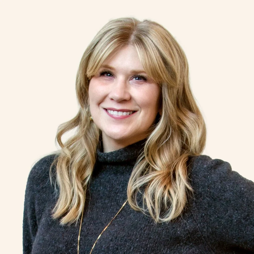 A woman with long wavy blonde hair wearing a black turtleneck sweater smiling at the camera against a plain light background.