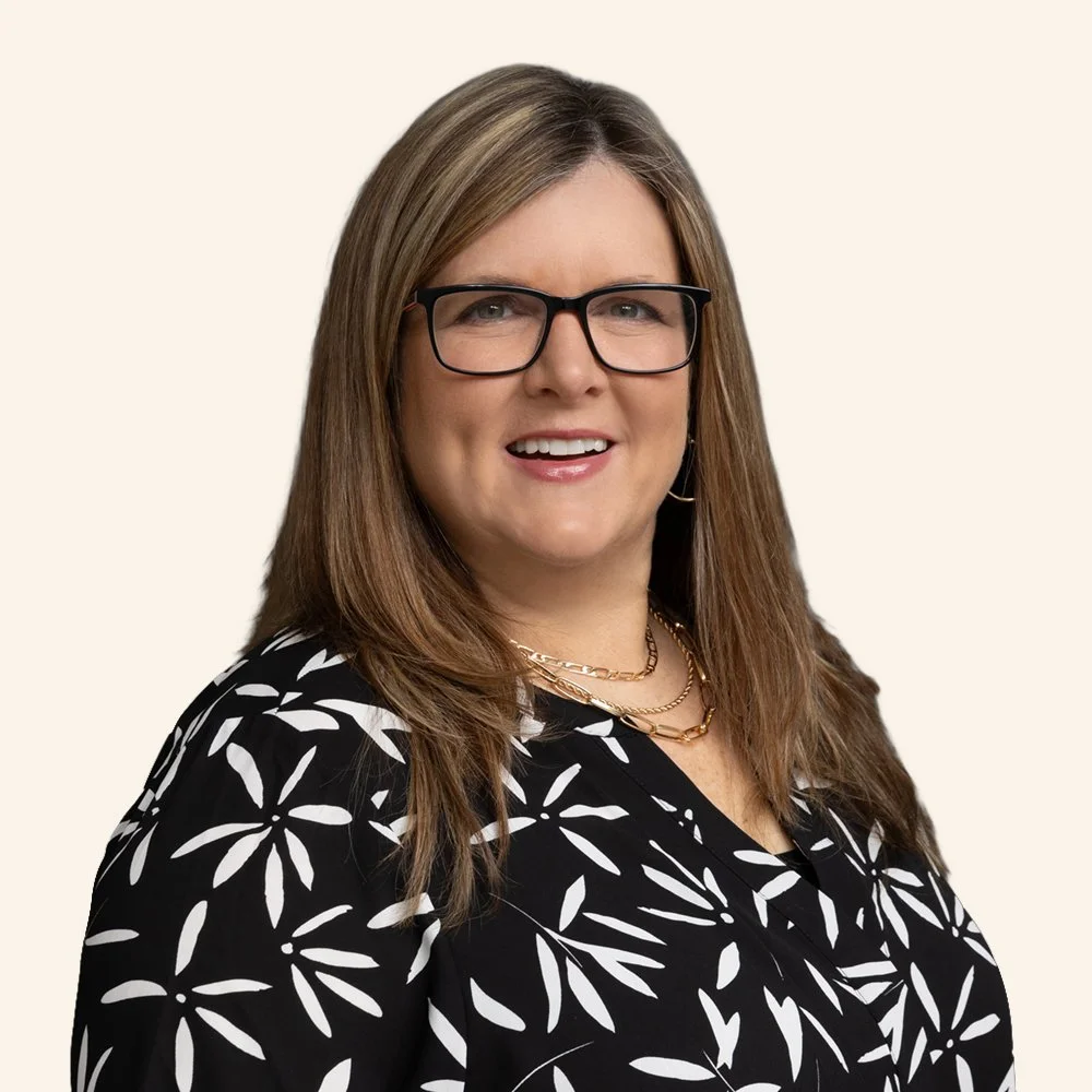 A woman with shoulder-length brown hair, wearing black-rimmed glasses, a black and white patterned blouse, and gold jewelry, smiling at the camera.