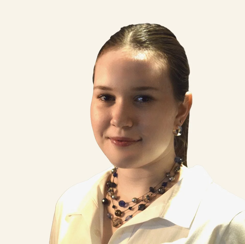 Young woman with brown hair tied back, wearing a white shirt, gold earrings, and layered pearl necklaces, smiling softly against a plain neutral background.