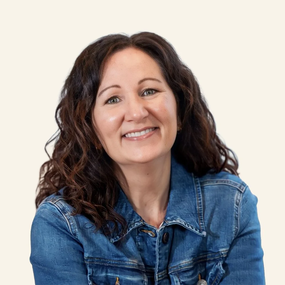 A woman with wavy brown hair smiling, wearing a denim jacket, against a plain background.