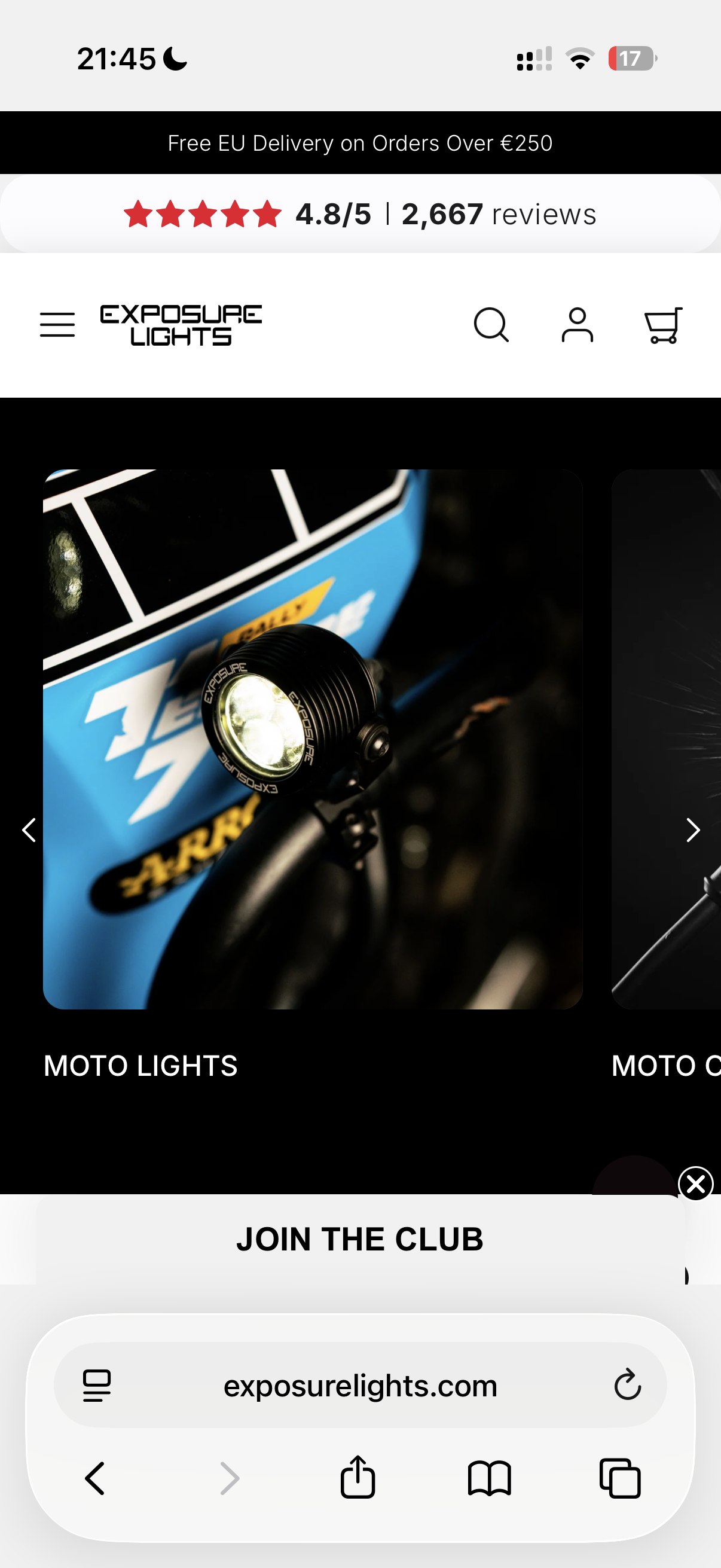 Close-up of a black motorcycle headlight with the brand name Exposure Lights, mounted on a bike with a blue frame. The background shows a blurred outdoor setting.