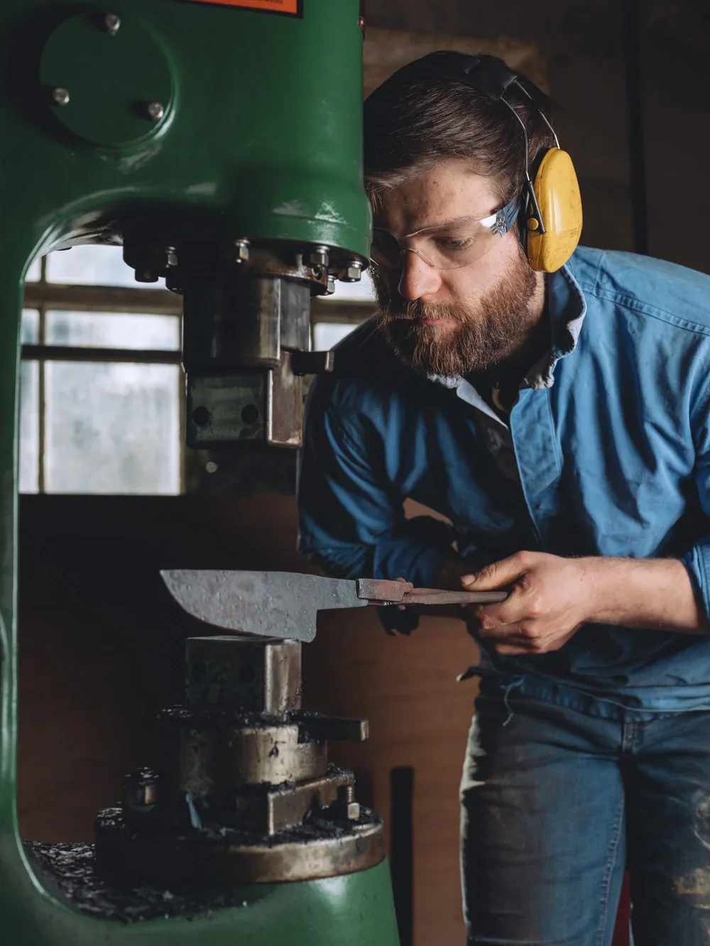A man with a beard, wearing yellow ear protection and safety glasses, operating a green industrial machine while examining a piece of metal.