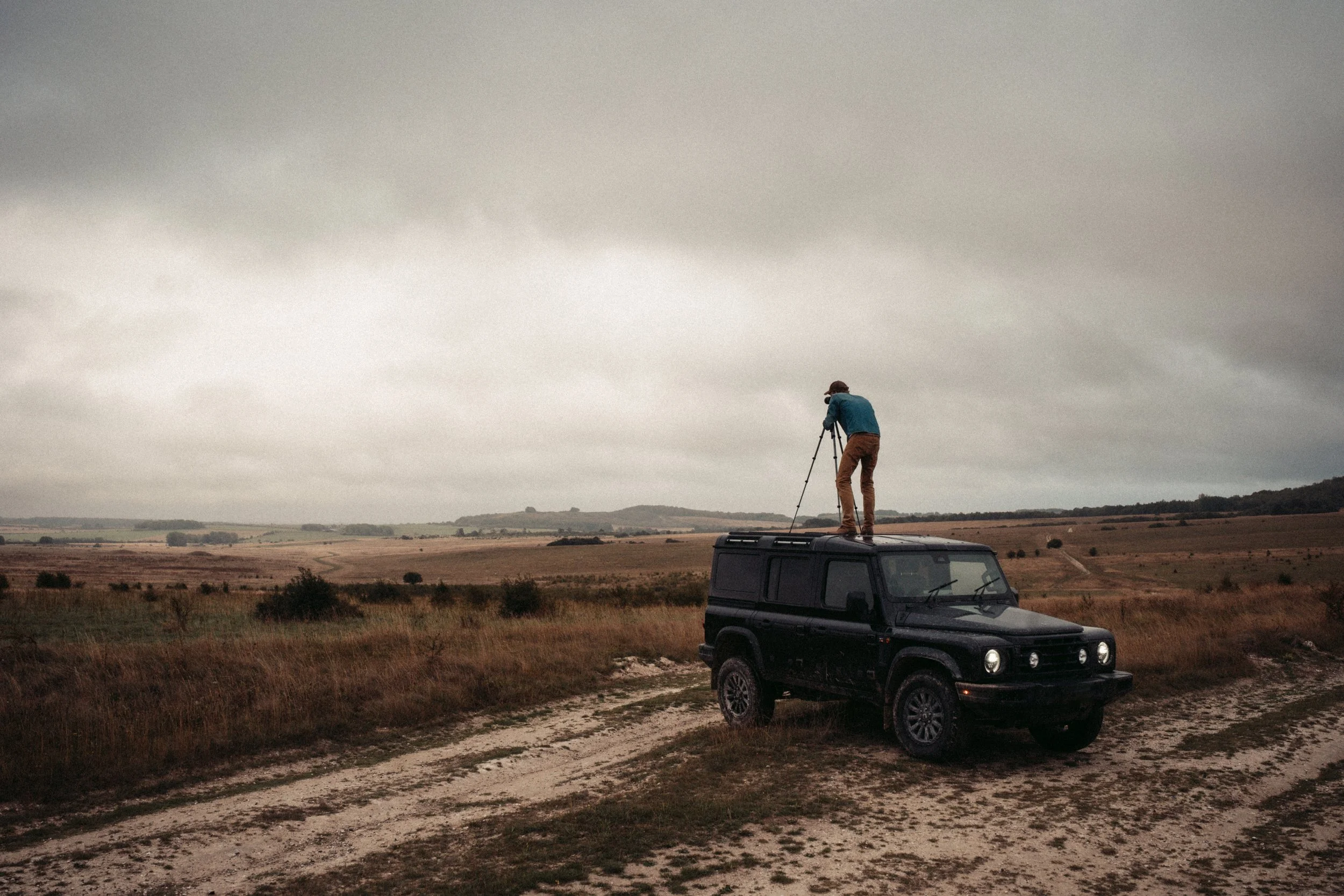 A person standing on top of a black SUV with a camera tripod, looking out over a rural landscape under cloudy skies.